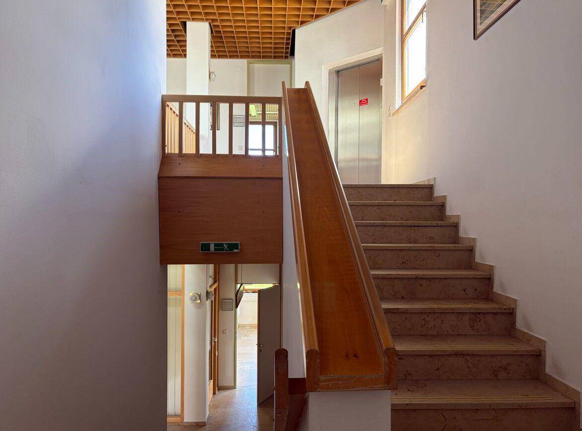 Treppenhaus mit Holzgeländer und einer Holzrutsche, die zum unteren Stockwerk führt, neben einem Aufzug.