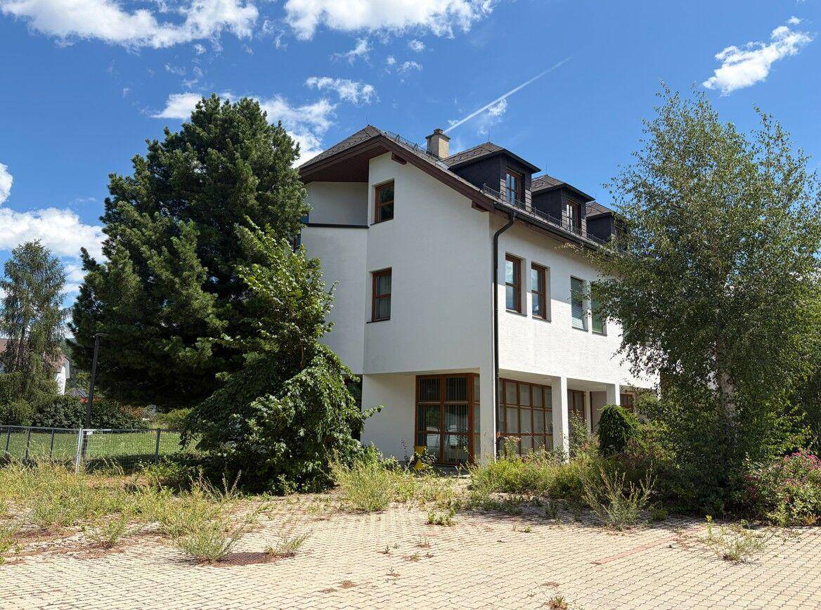 Ansicht des Gebäudes von der Seite mit weißer Fassade, vielen Fenstern und umgebender Vegetation.