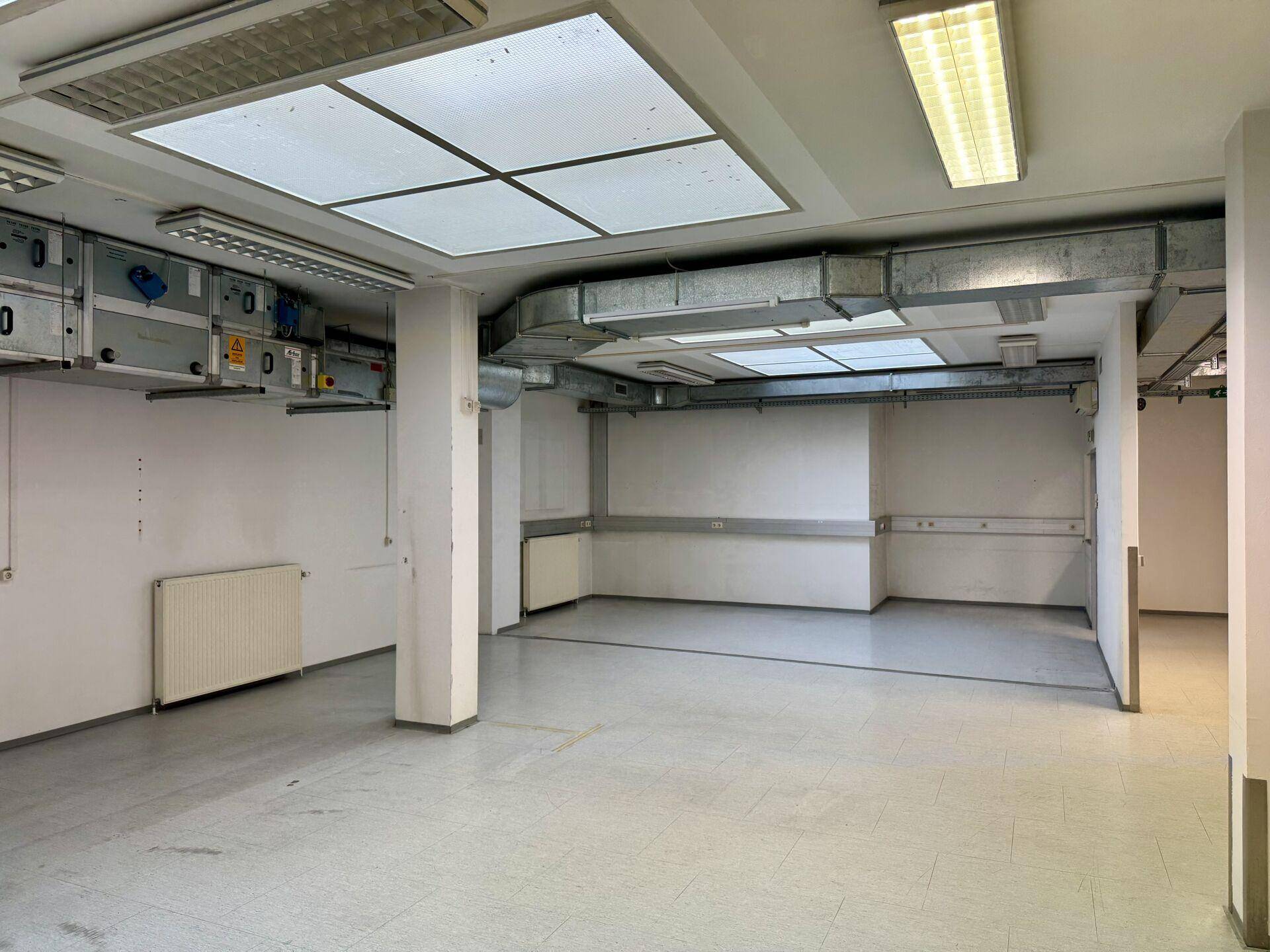 Großer, leerer Gewerberaum mit Fliesenboden, Heizkörpern und sichtbarer Lüftungsanlage an der Decke.