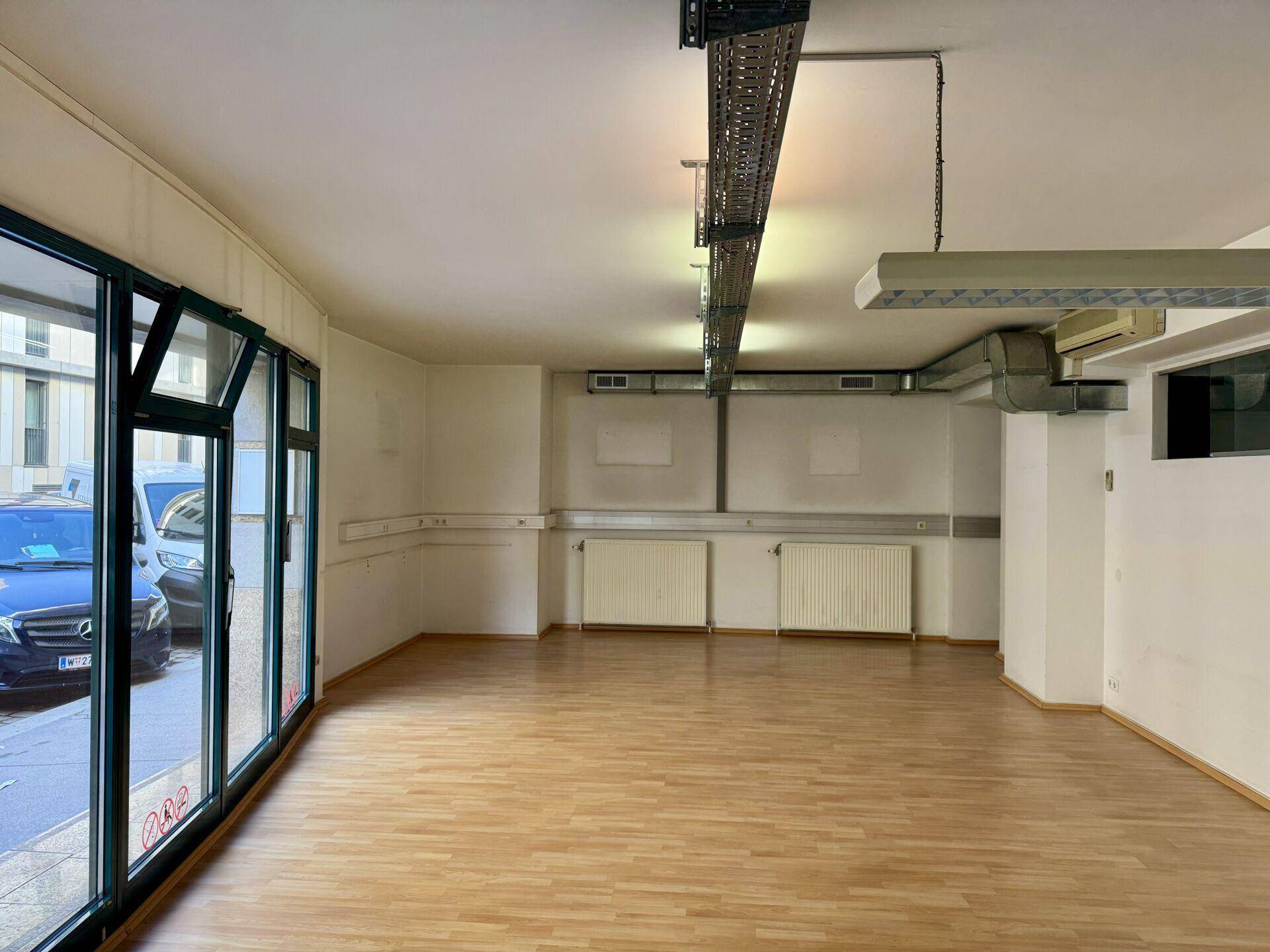 Leerer Gewerberaum mit großen Schaufenstern, Laminatboden und sichtbaren Heizkörpern.