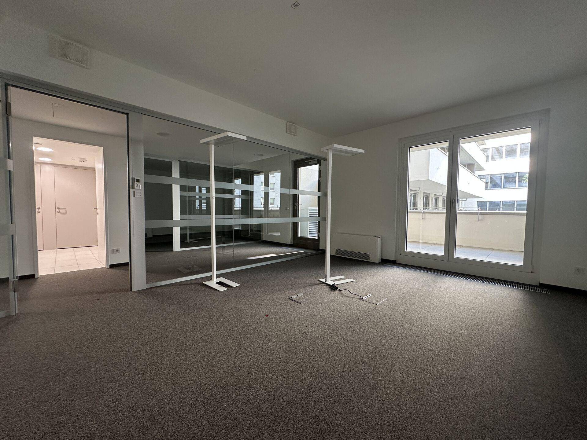 Helles Bürozimmer mit Glaswänden, Teppichboden und Zugang zu einem Balkon oder einer Terrasse.