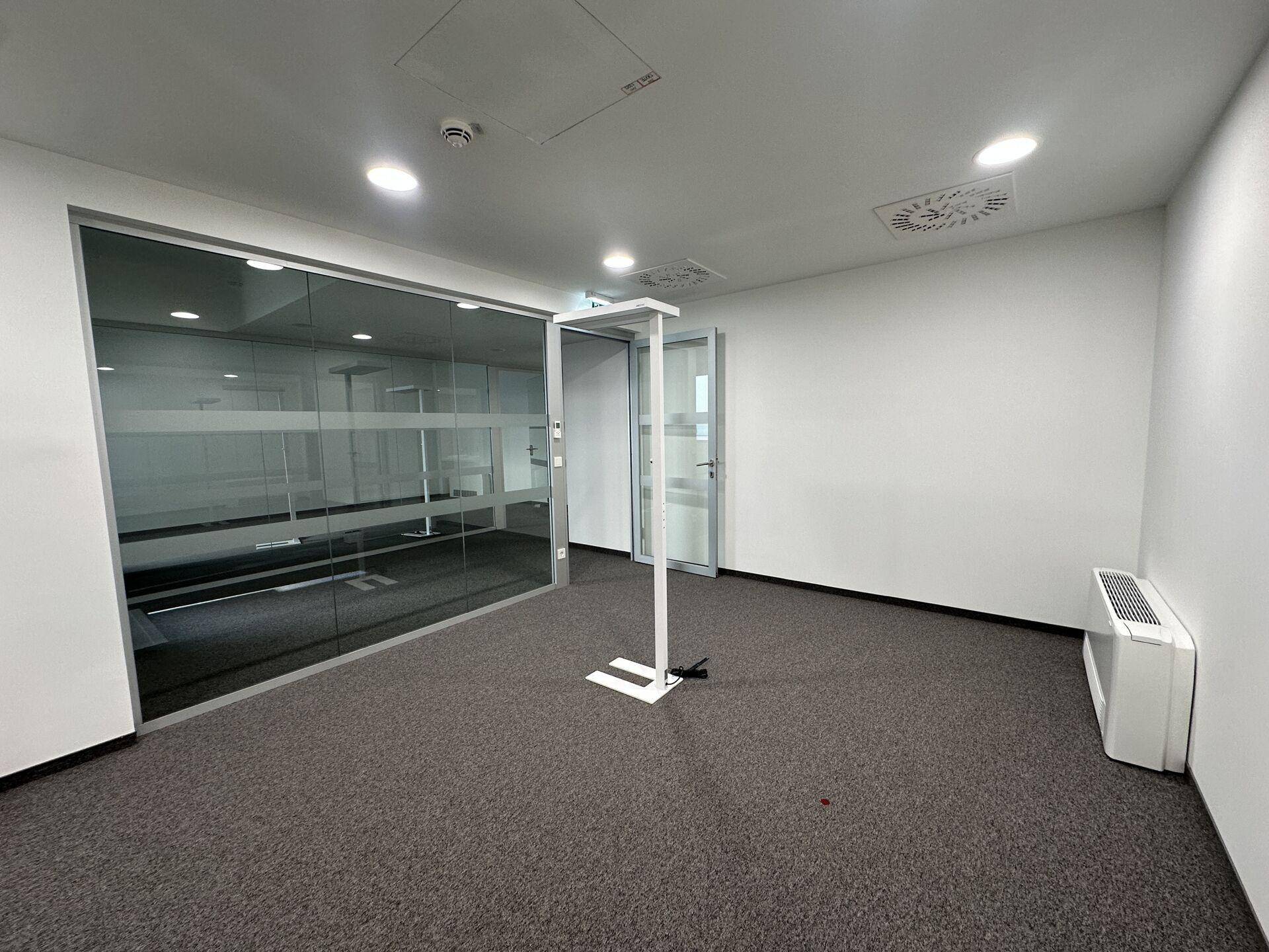 Modernes Bürozimmer mit Glaswänden, Teppichboden und sichtbarer Lüftungsanlage an der Decke.