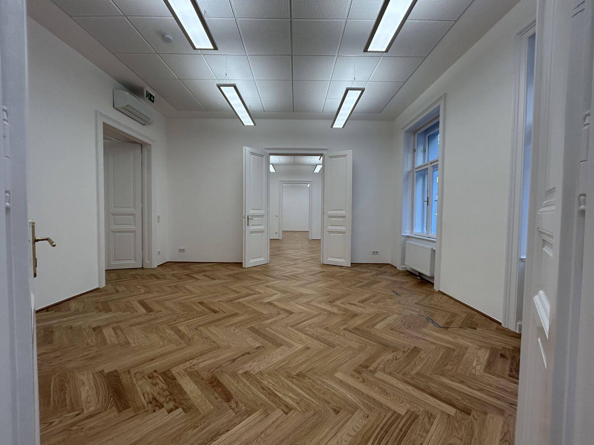 Geräumiges leeres Büro mit Fischgrätparkettboden und mehreren Türen, die zu weiteren Räumen führen.