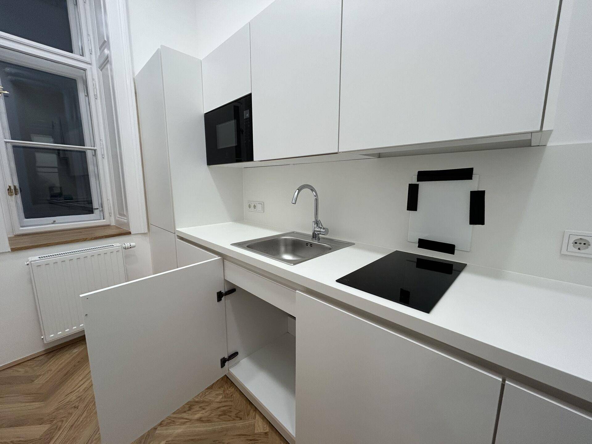 Moderne weiße Teeküche mit Induktionskochfeld, Spüle und eingebauter Mikrowelle, hell durch ein Fenster.