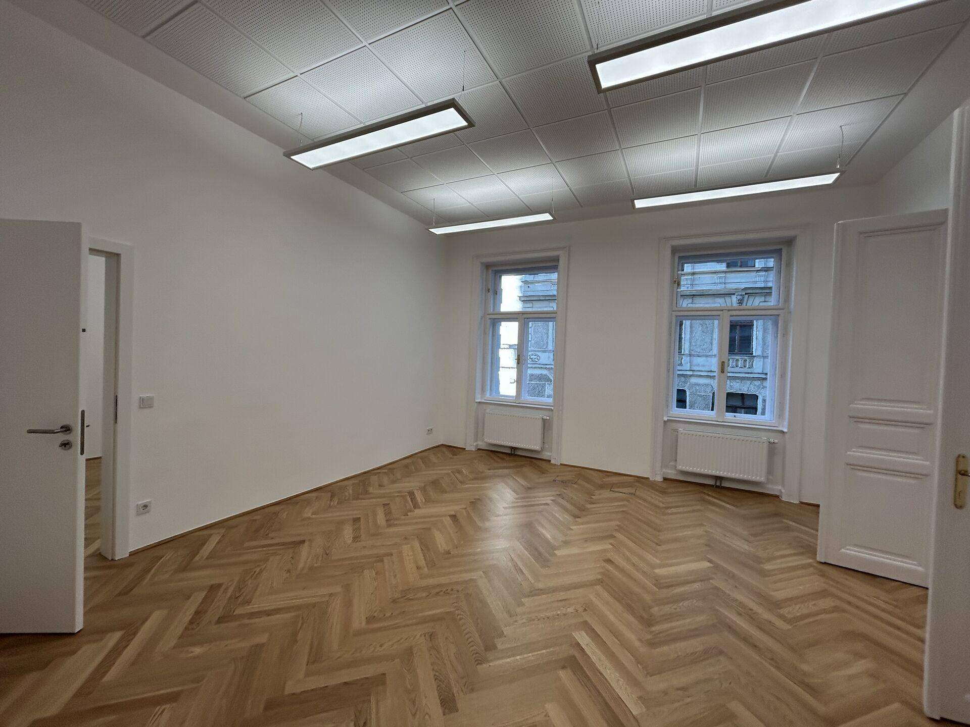 Leeres Büro mit Fischgrätparkettboden, weißen Wänden und zwei großen Fenstern, bereit zur Einrichtung.