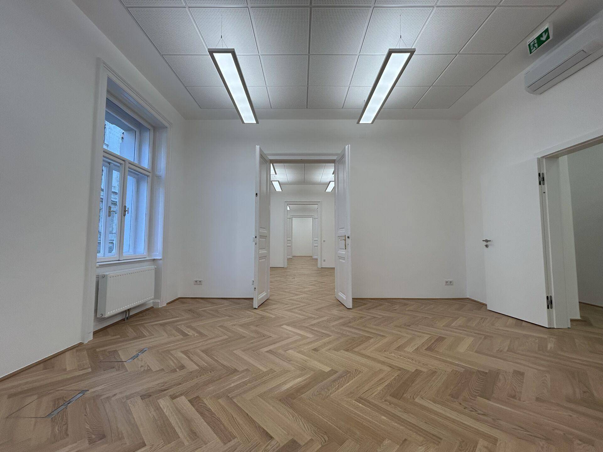 Geräumiges leeres Büro mit Fischgrätparkettboden und mehreren Türen, die zu weiteren Räumen führen.