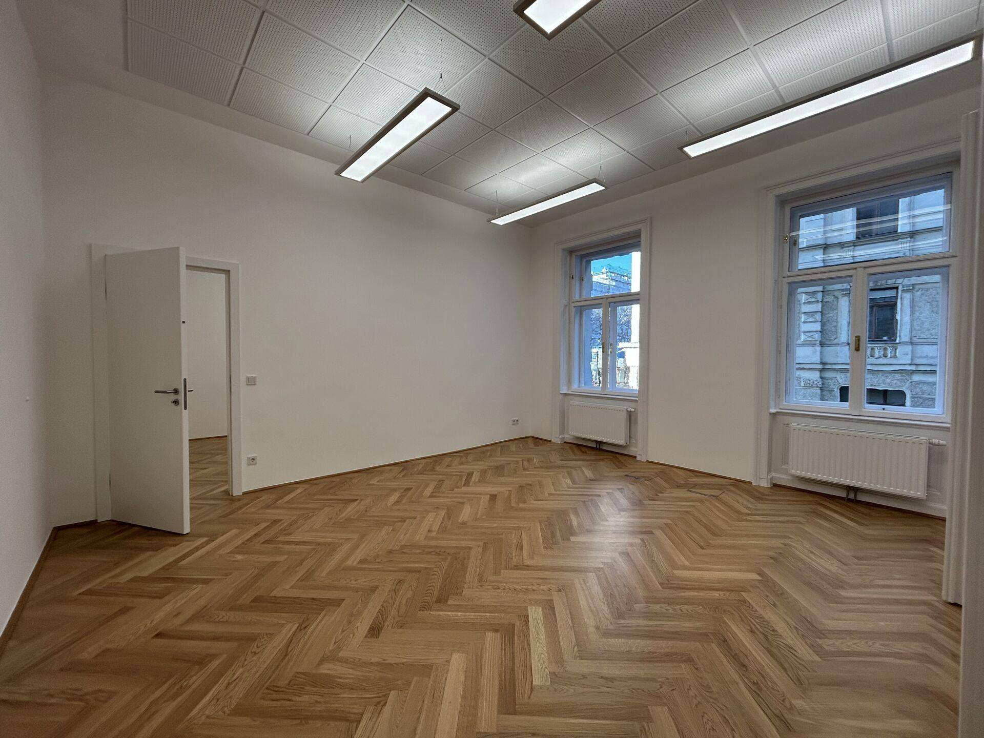 Leeres Büro mit Fischgrätparkettboden, weißen Wänden und zwei großen Fenstern, bereit zur Einrichtung.