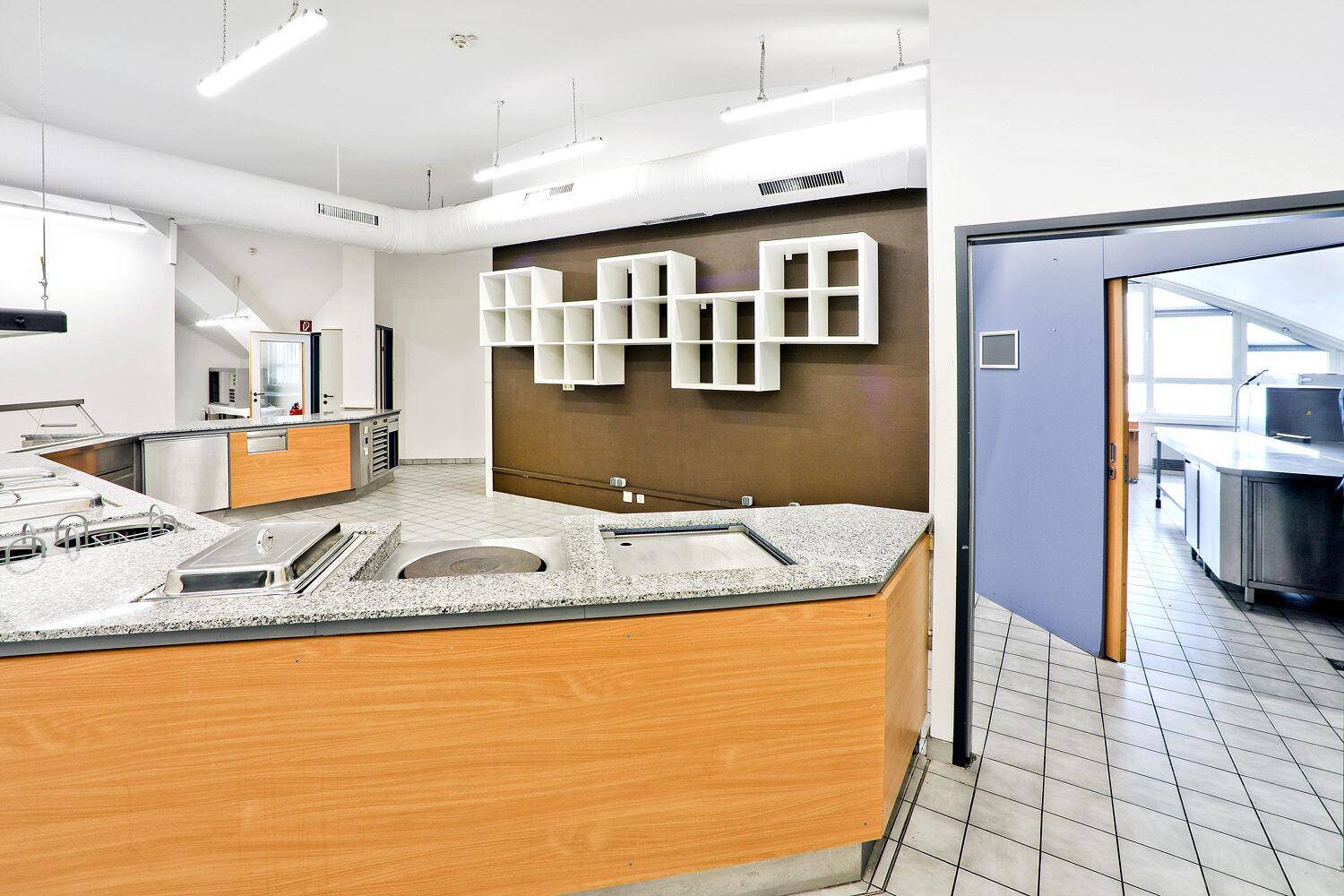 Kantinenküche mit Servicetheke, Holzverkleidung und Regalen an einer braunen Akzentwand.