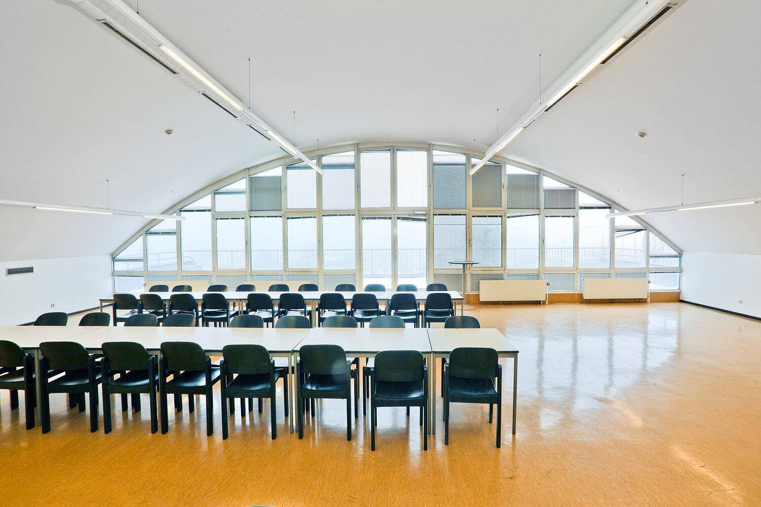 Großer Konferenzraum mit vielen Tischen und Stühlen, gewölbter Fensterfront und hellem Boden.
