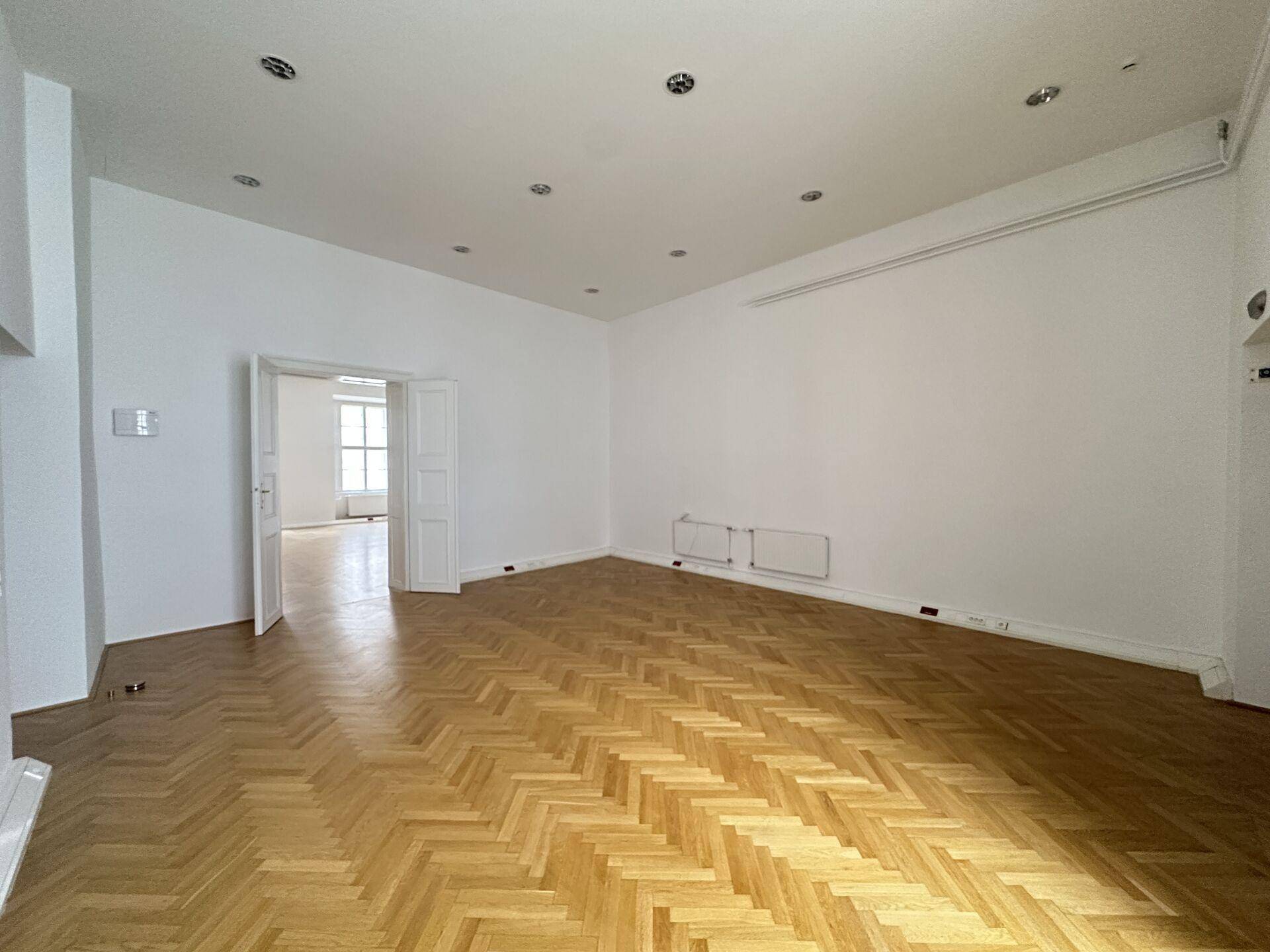 Heller, leerer Raum mit Fischgrätparkett, weißen Wänden und offenem Durchgang zu einem weiteren Zimmer.