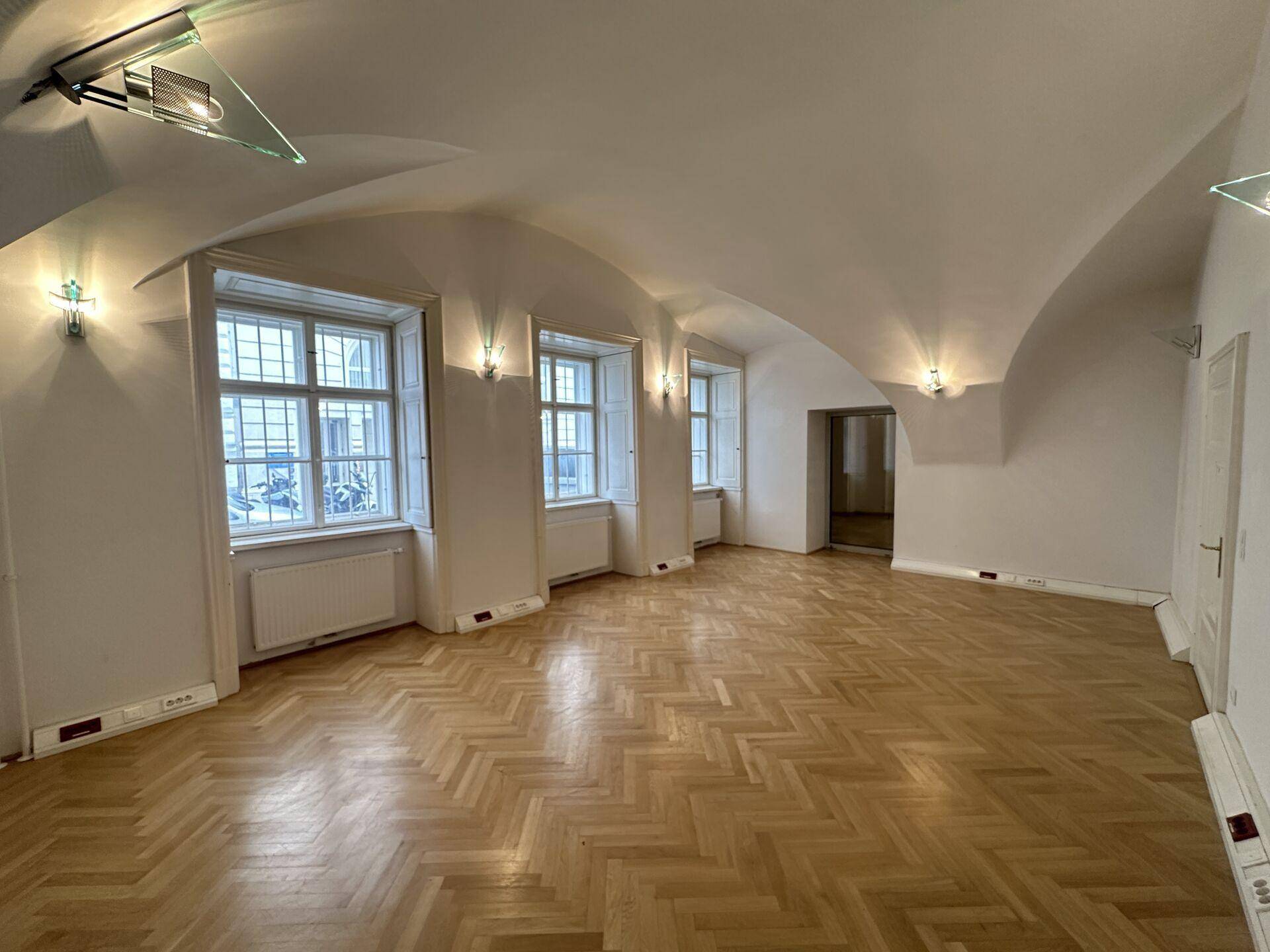 Eleganter Raum mit gewölbter Decke, Fischgrätparkett und drei Fenstern mit dekorativen Wandleuchten.