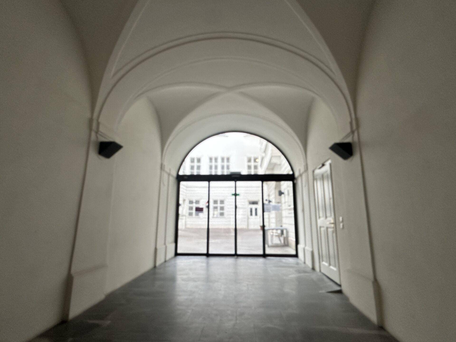 Gewölbter Durchgang mit hohen Decken und einer modernen Glastür, die zu einem Innenhof führt.