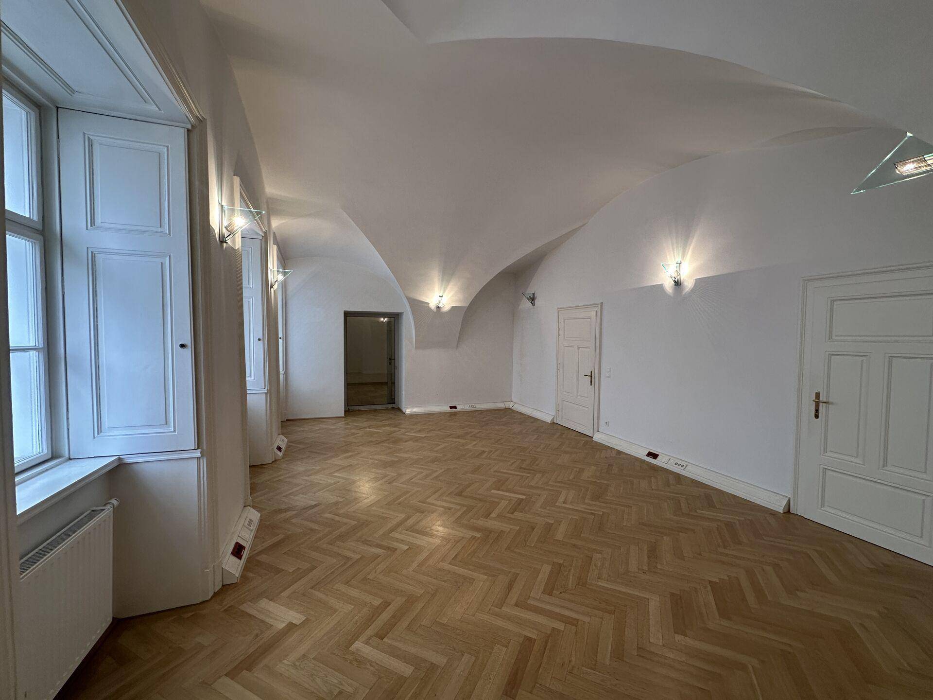 Geräumiger Raum mit eleganter Gewölbedecke, Fischgrätparkett und Fenstern mit Wandbeleuchtung.