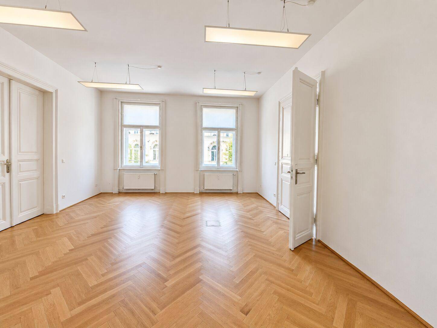 Geräumiger Büroraum mit klassischem Fischgrätparkett und zwei großen Fenstern.