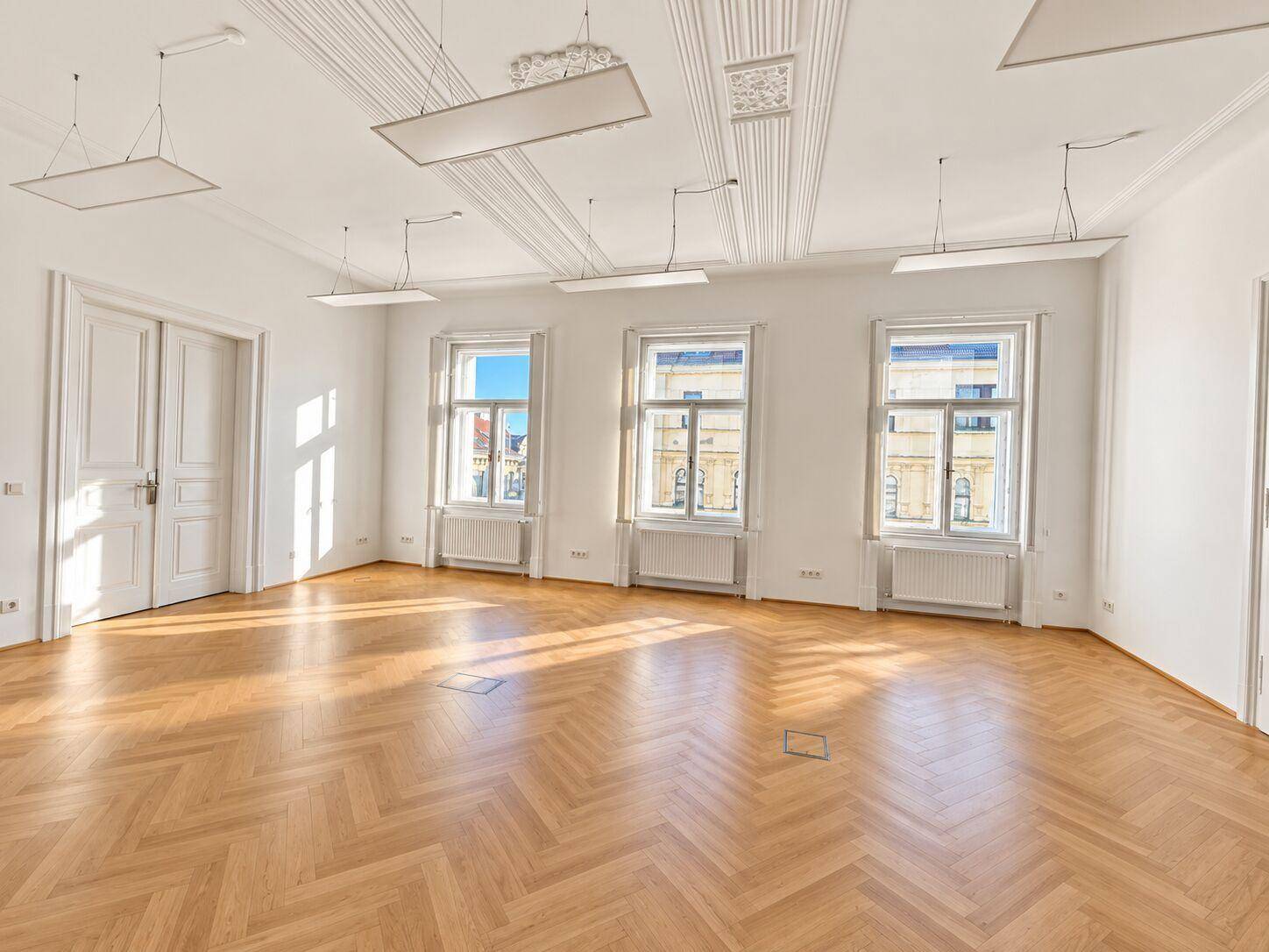 Großer, leerer Raum mit Fischgrätparkettboden, hohen Decken und drei großen Fenstern, ideal für ein Büro.