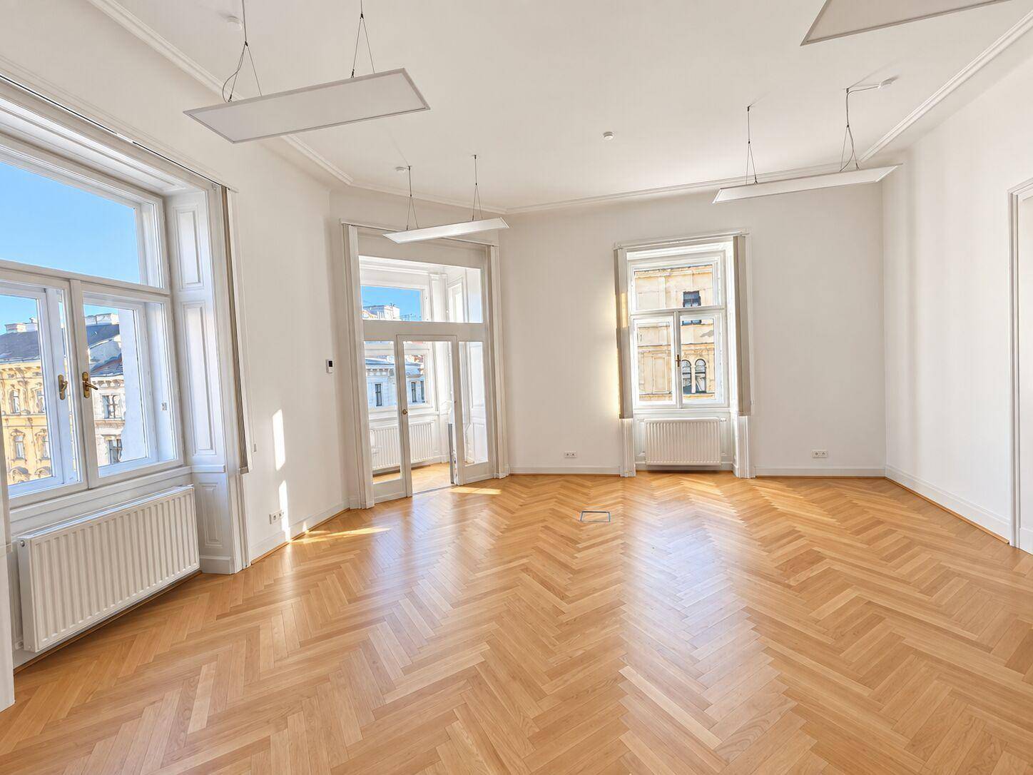Heller Büroraum mit Fischgrätparkett, hohen Fenstern und Blick auf die Stadt.
