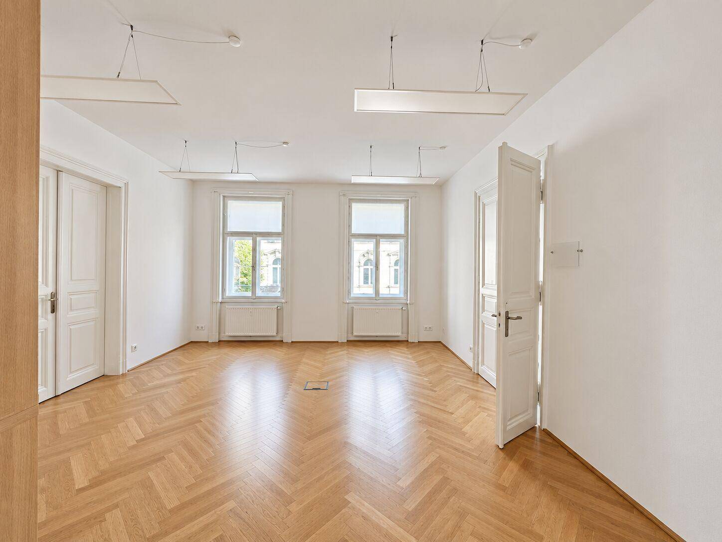 Leerer Büroraum mit Fischgrätparkett, weißen Wänden und zwei Fenstern mit Rollos.