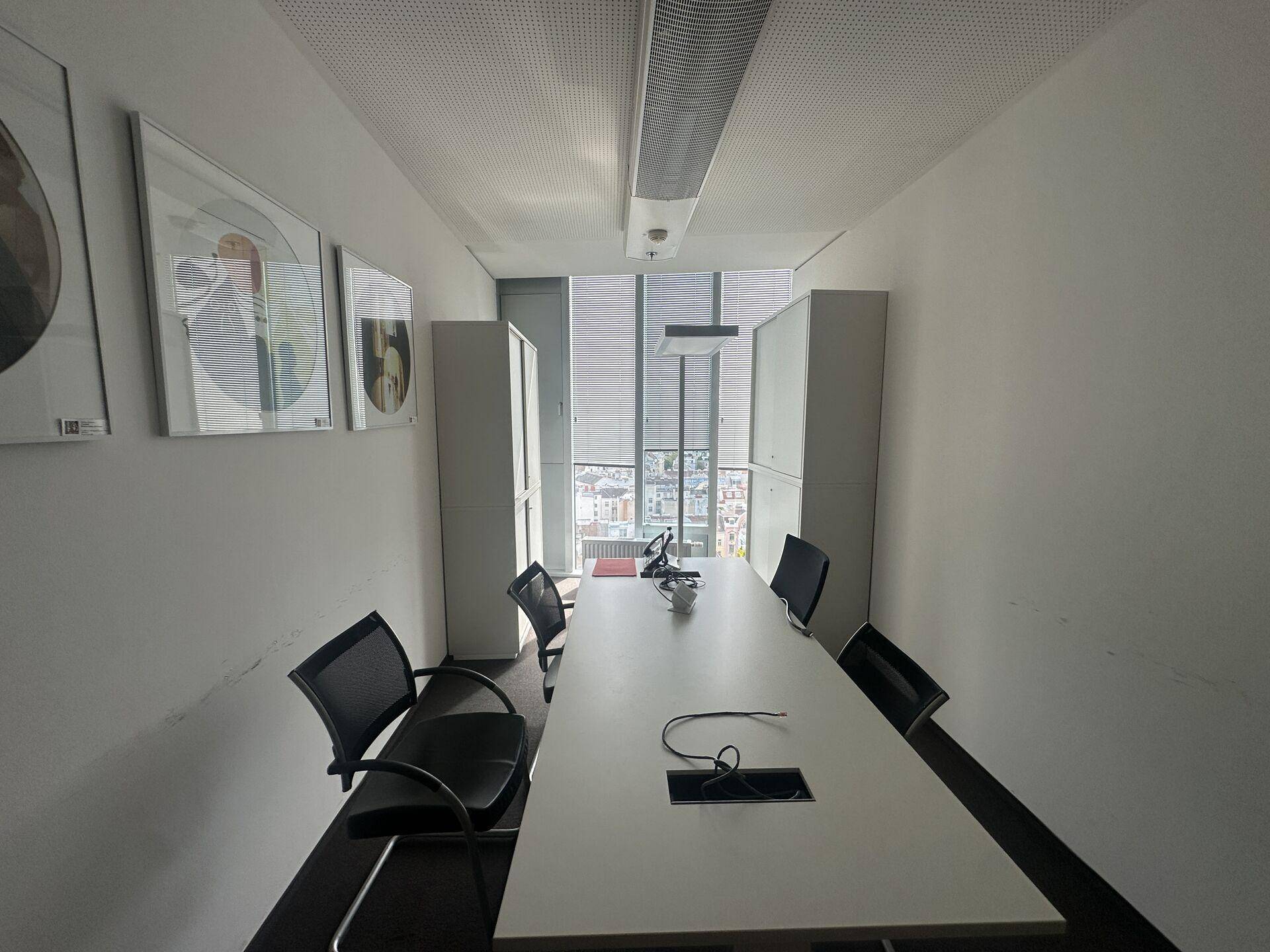 Schmaler Konferenzraum mit langem weißen Tisch, Bürostühlen und einem Fenster am Ende, das Tageslicht spendet.