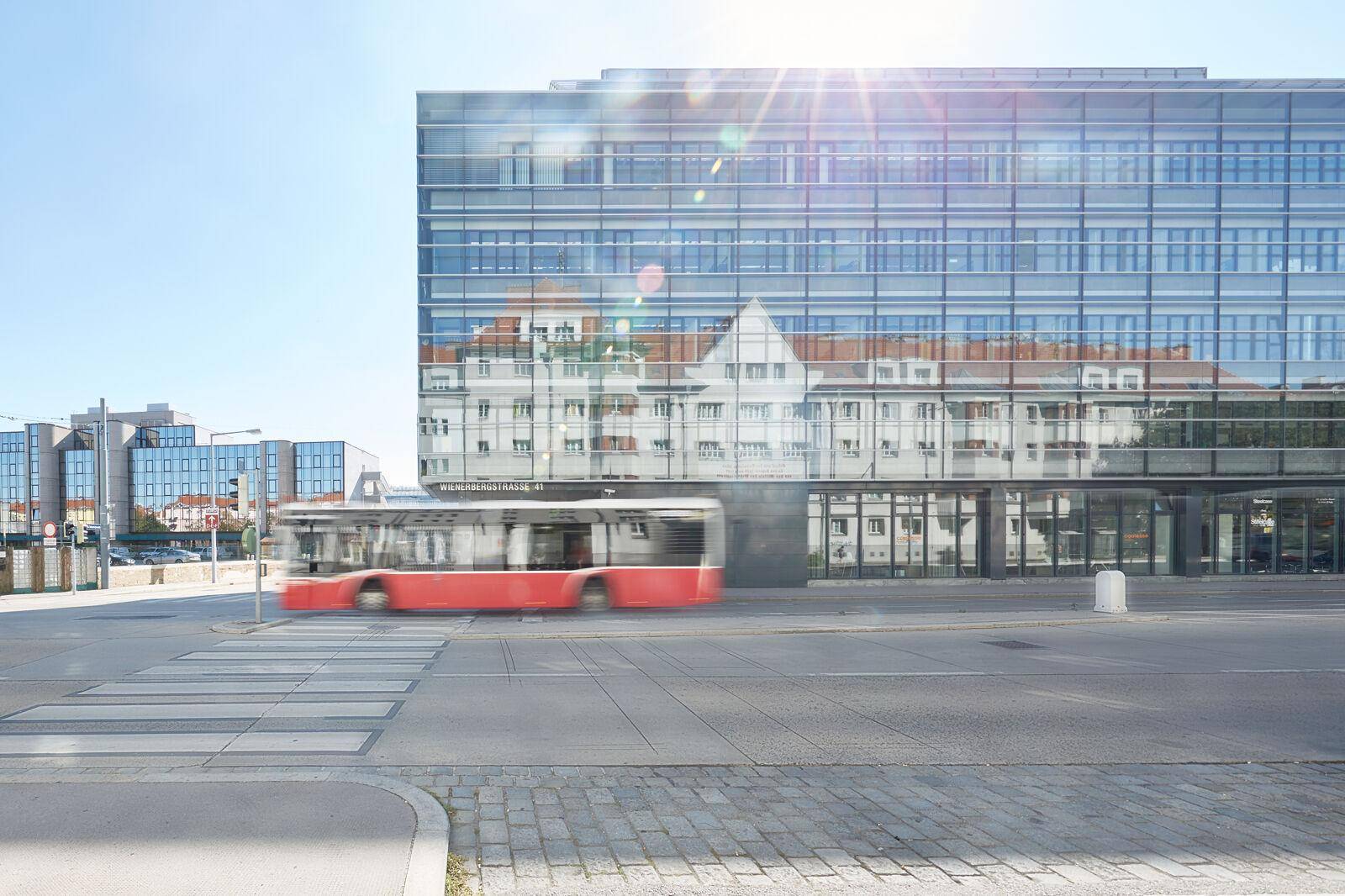 Moderne Glasfassade eines Bürogebäudes spiegelt die Umgebung wider, während ein Bus vorbeifährt.