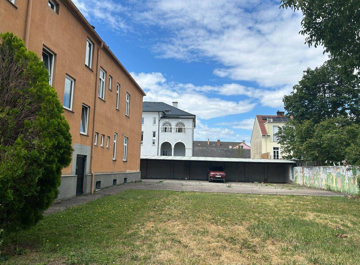 Seitenansicht des Gebäudes mit einer Grünfläche und einem überdachten Parkplatz im Hintergrund.