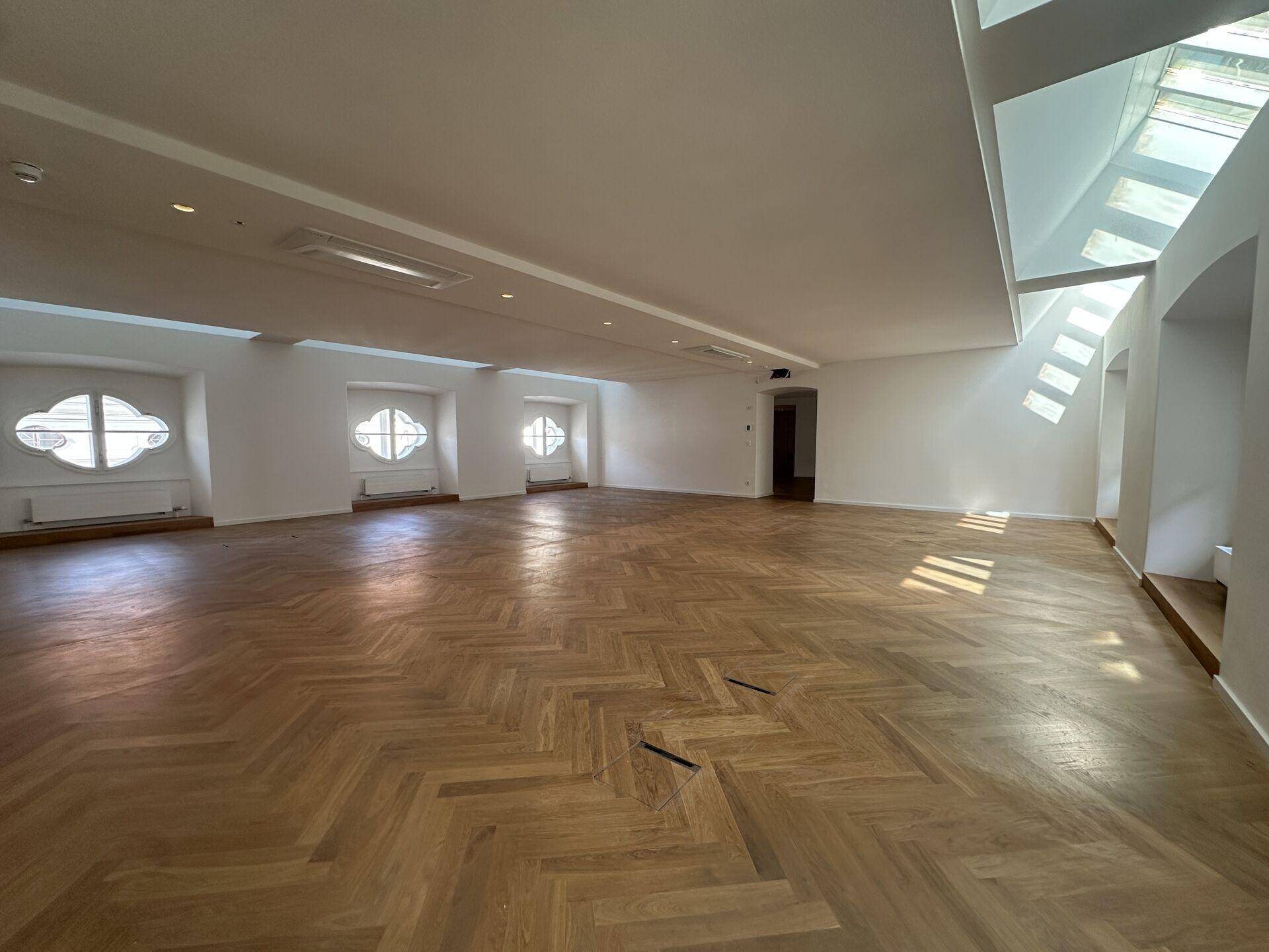Geräumiger, leerer Raum mit Fischgrätparkett, hohen Decken und ungewöhnlichen Fenstern, die viel Licht hereinlassen.