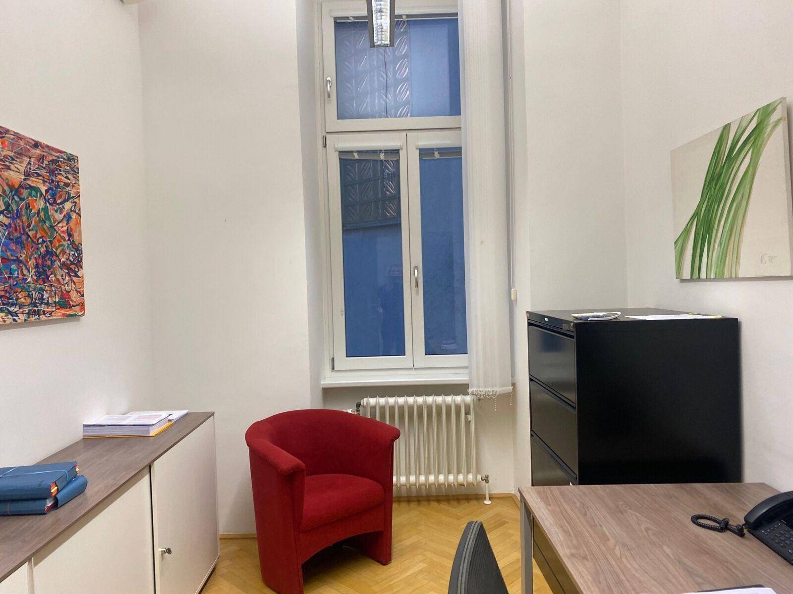 Kleines Büro mit rotem Sessel, Aktenschrank und Schreibtisch, hell durch ein großes Fenster.