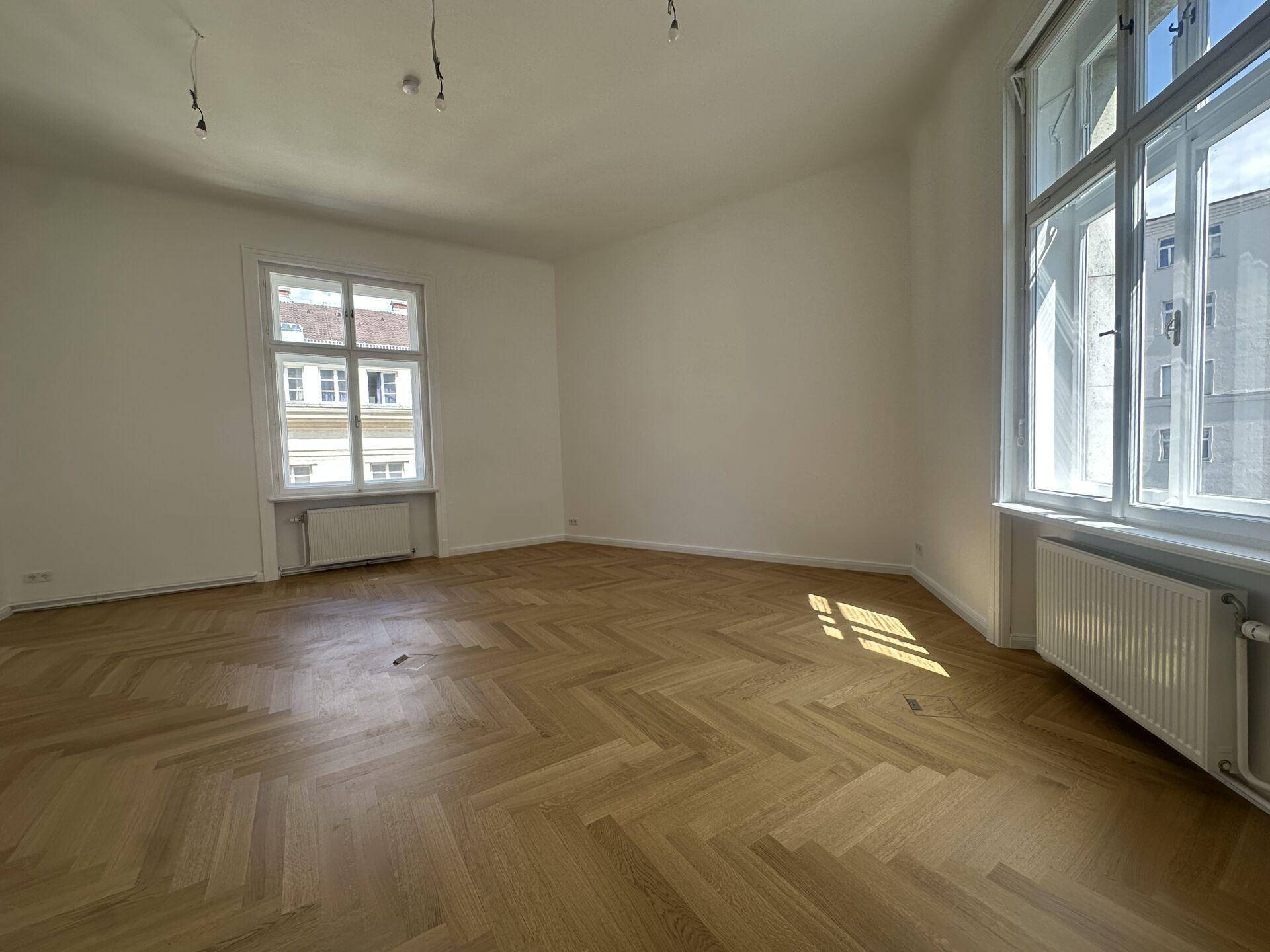 Geräumiger Büroraum mit hellem Fischgrätparkett und großen Fenstern, die viel Tageslicht hereinlassen.