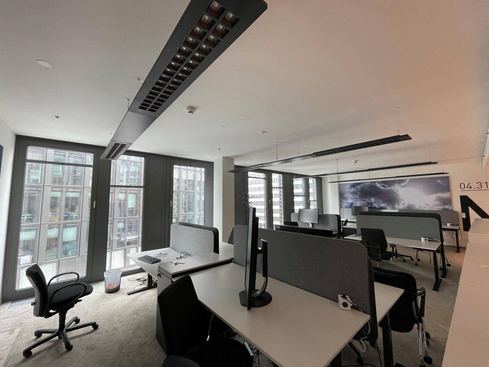 Offener Bürobereich mit mehreren Arbeitsplätzen, großen Fenstern und Blick auf andere Gebäude.