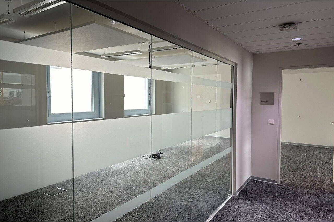 Moderner Bürobereich mit einer Glaswand, die einen weiteren Raum mit Fenstern und Teppichboden abtrennt.