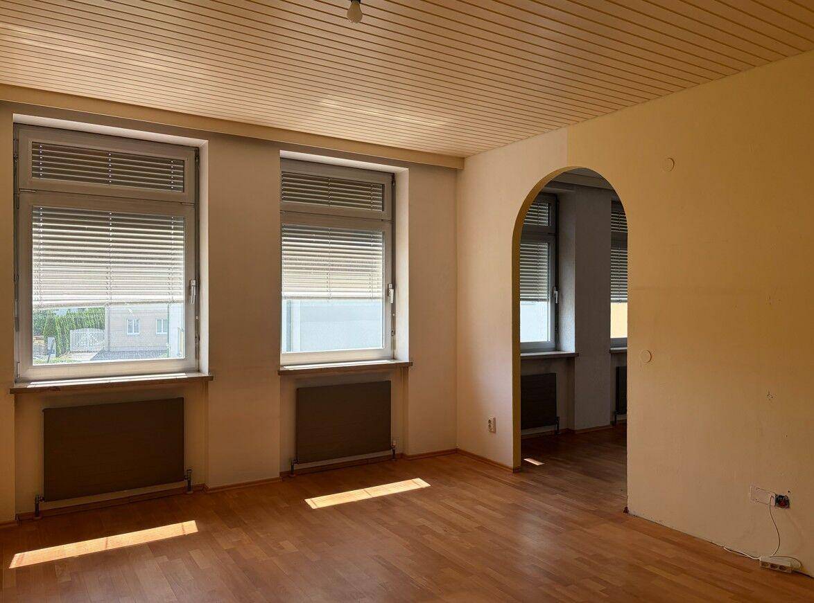 Leerer Wohnraum mit Holzboden, zwei Fenstern und einem Rundbogen zu einem weiteren Zimmer.