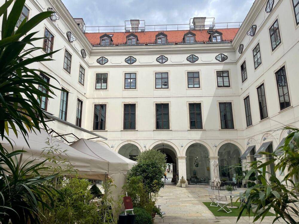 Heller Innenhof mit Arkaden, Pflanzen und Sitzgelegenheiten, umgeben von historischen Gebäudefassaden.