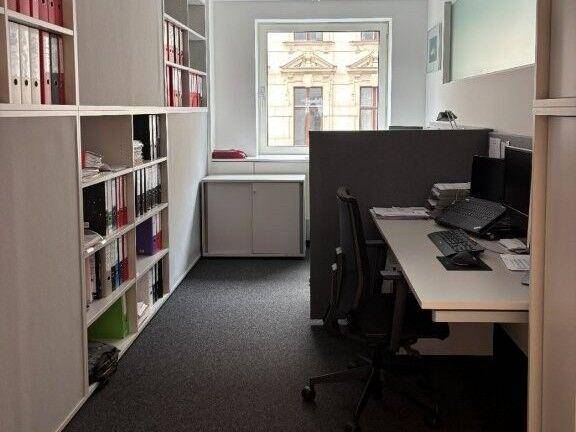 Kleines Büro mit einem Arbeitsplatz, Regalen voller Akten und einem Fenster mit Blick auf ein historisches Gebäude.