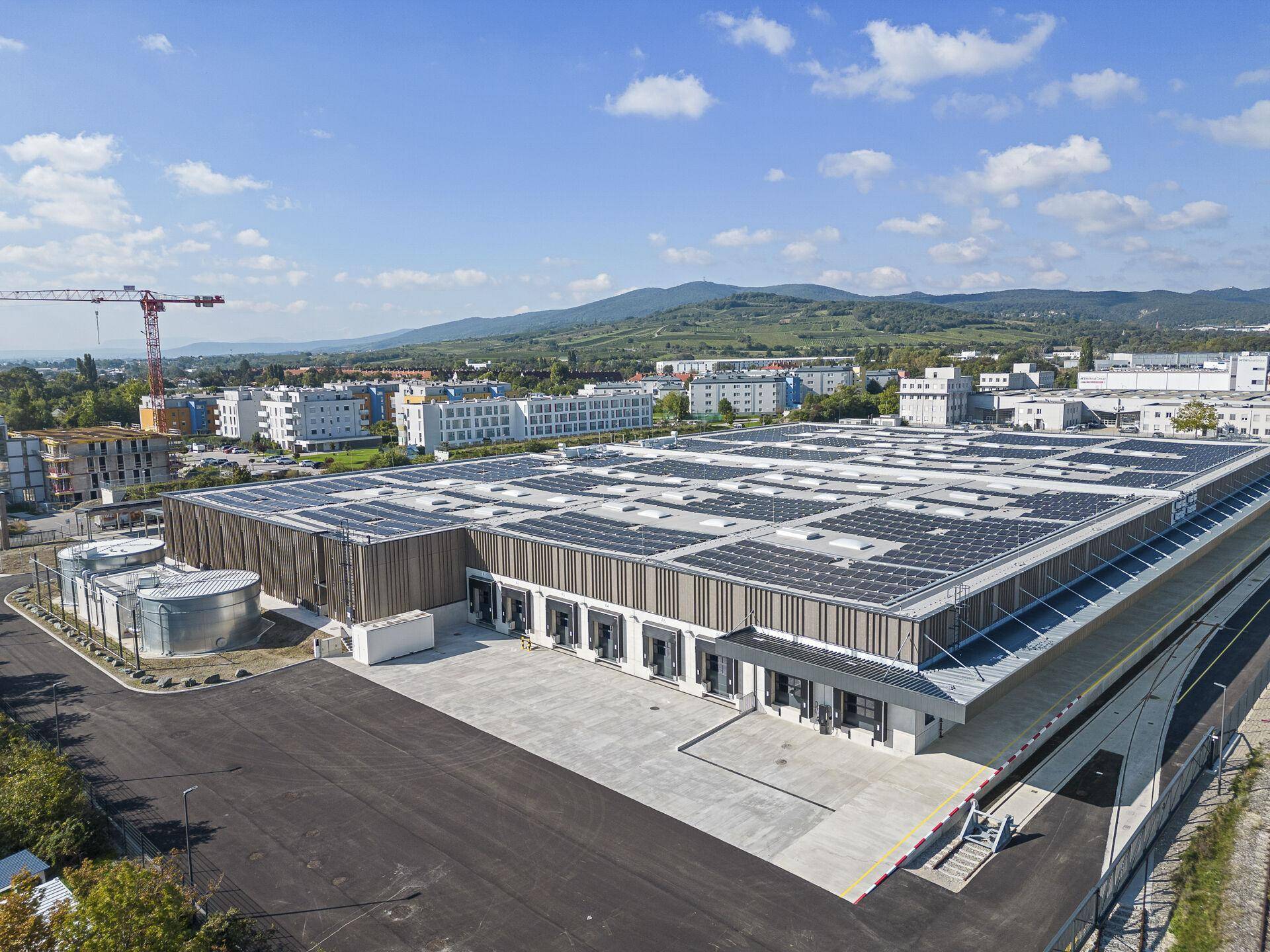 Panoramische Luftaufnahme eines großen Industriegebäudes mit Solaranlage, umgeben von Wohngebäuden und Bergen im Hintergrund.