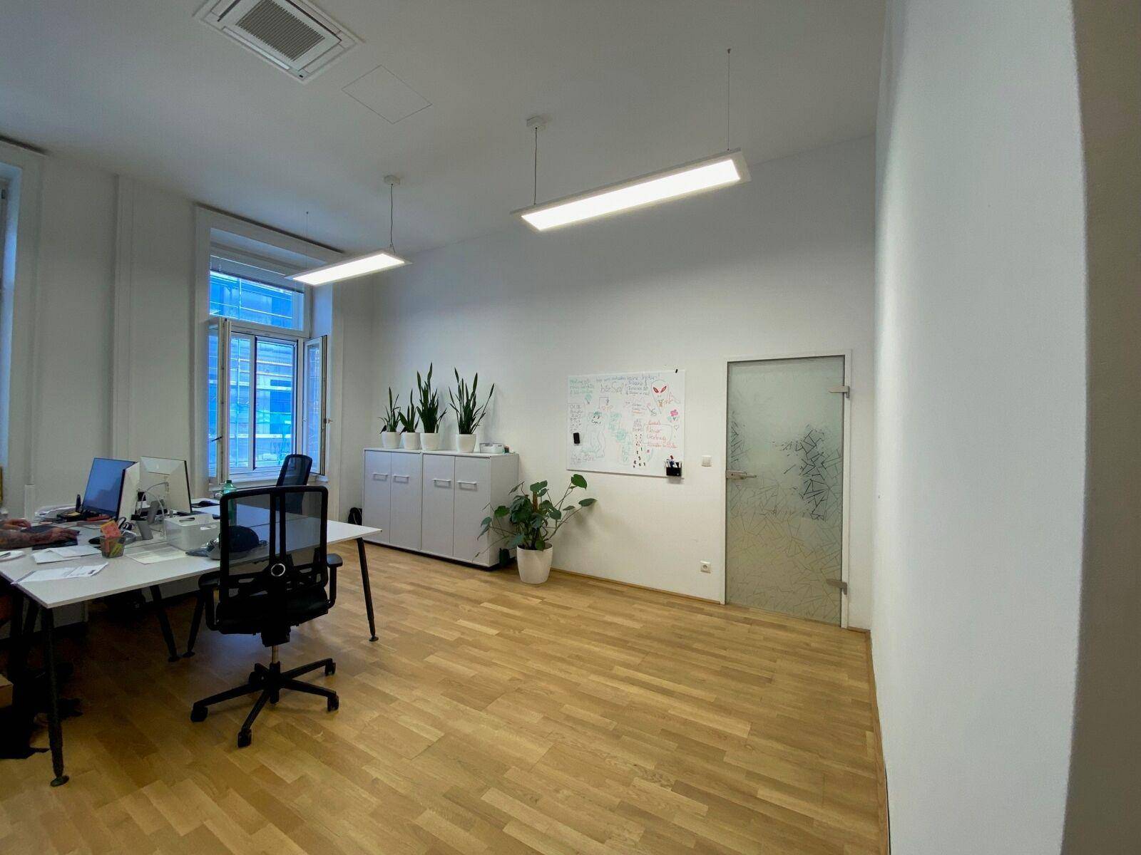 Büroraum mit Holzparkett, Schreibtischen, Whiteboard und einer Milchglastür.