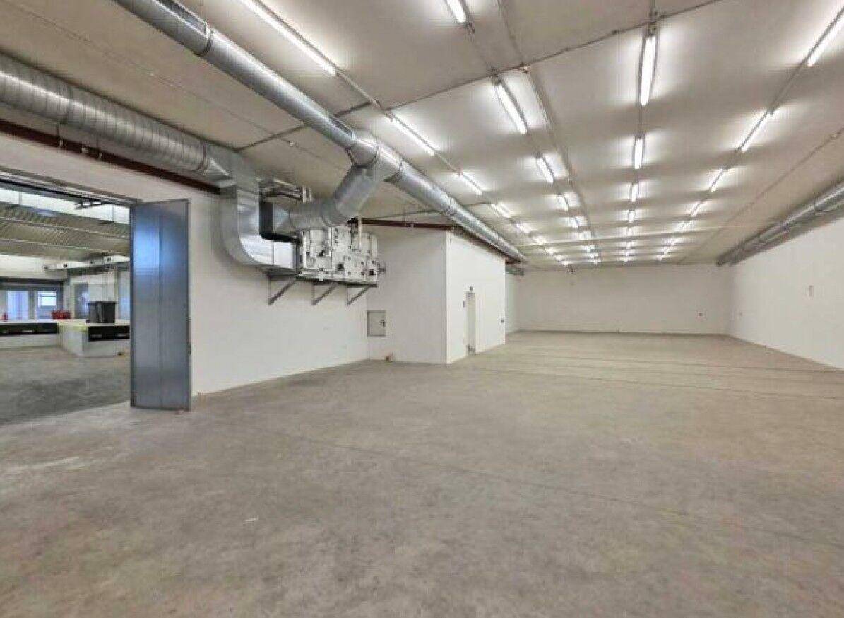 Geräumiger, leerer Gewerberaum mit Betonboden, sichtbaren Lüftungsrohren und vielen Deckenleuchten.