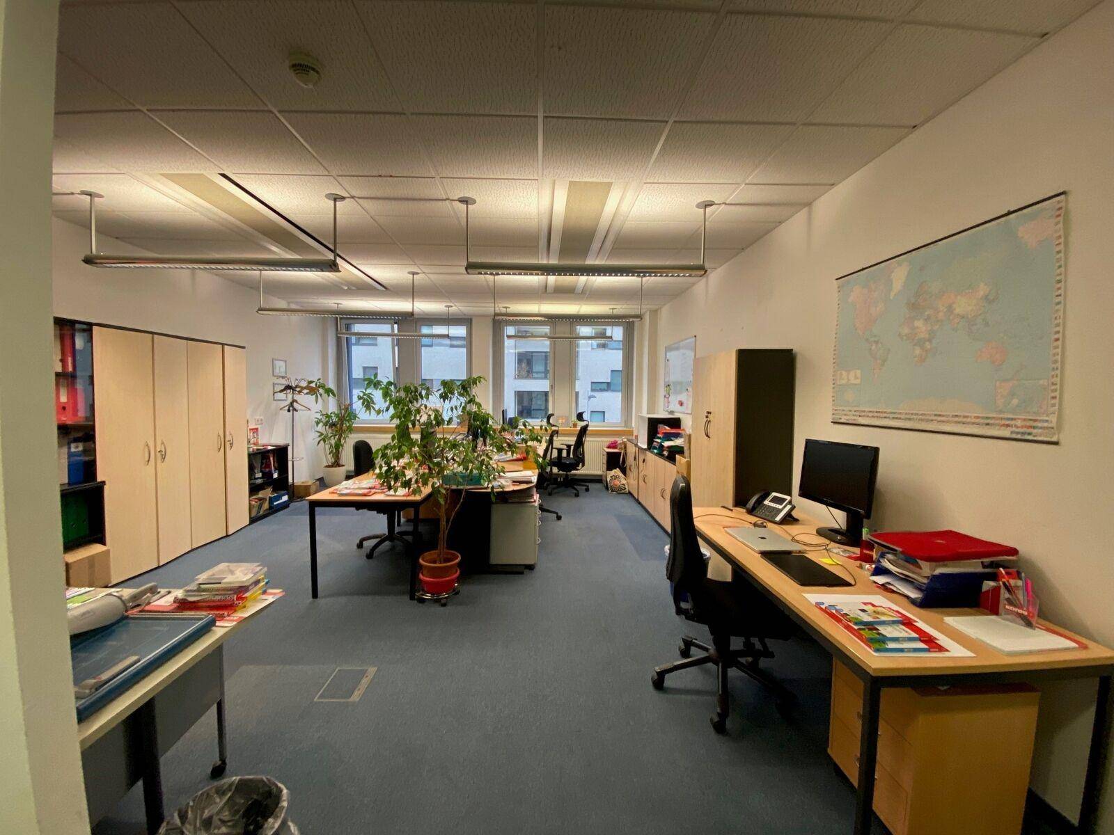 Großraumbüro mit mehreren Arbeitsplätzen, großen Fenstern und vielen Pflanzen für eine angenehme Atmosphäre.