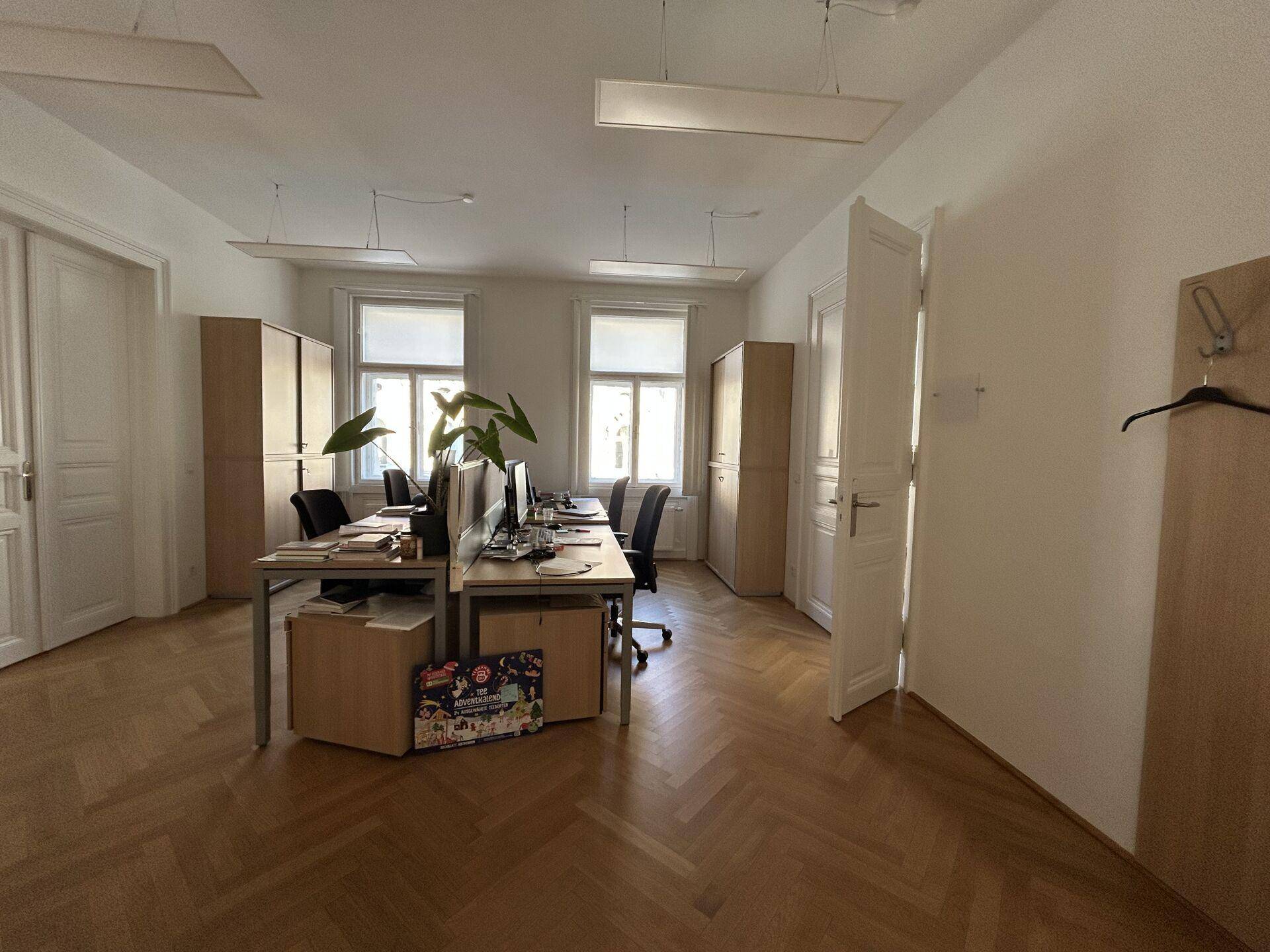 Helles Büro mit mehreren Arbeitsplätzen, Holzschränken und Fischgrätparkettboden, gut beleuchtet durch große Fenster.