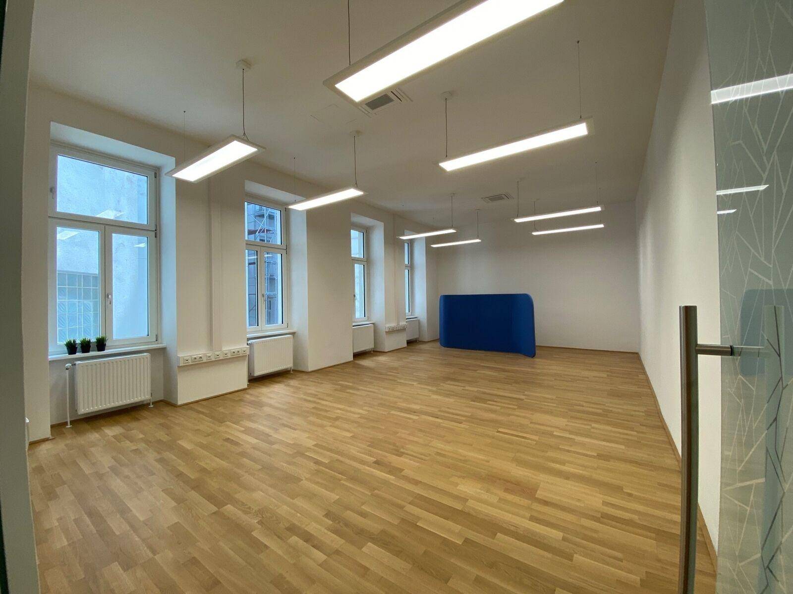 Geräumiger, leerer Büroraum mit Holzparkett, großen Fenstern und modernen Deckenleuchten.