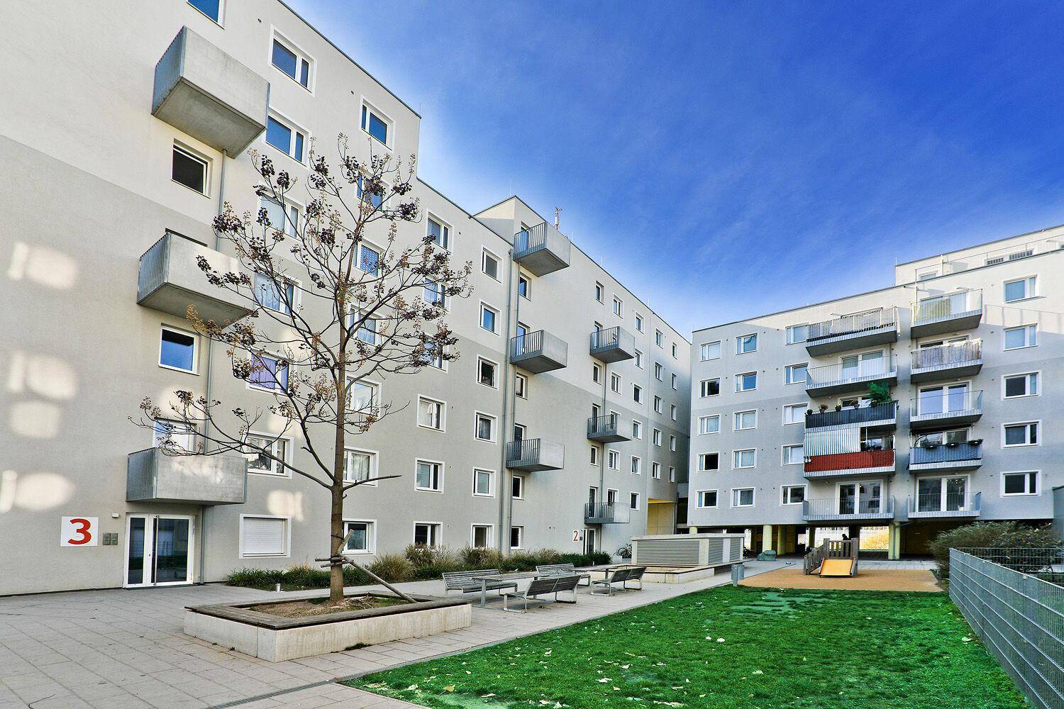 Moderner Innenhof mit Spielplatz und Sitzbänken zwischen den Wohngebäuden unter blauem Himmel.