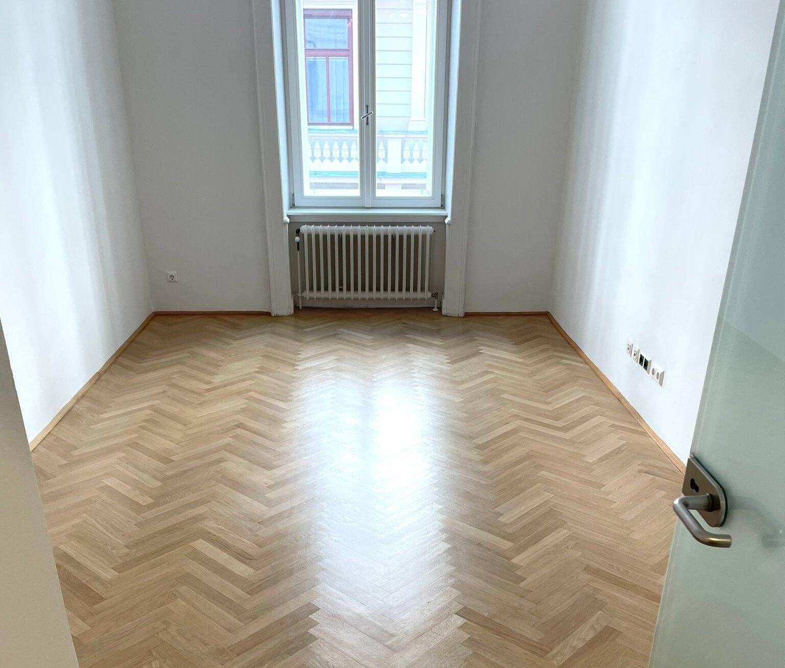Leerer Raum mit Fischgrätparkett, weißen Wänden und einem Fenster mit Heizkörper.