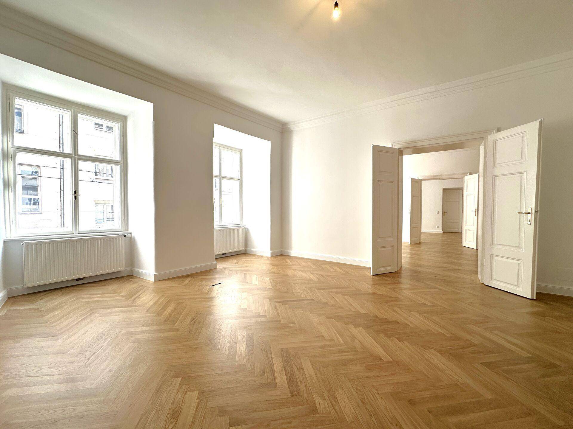 Geräumiges Wohnzimmer mit Fischgrätparkett, hohen Decken und großen Fenstern.