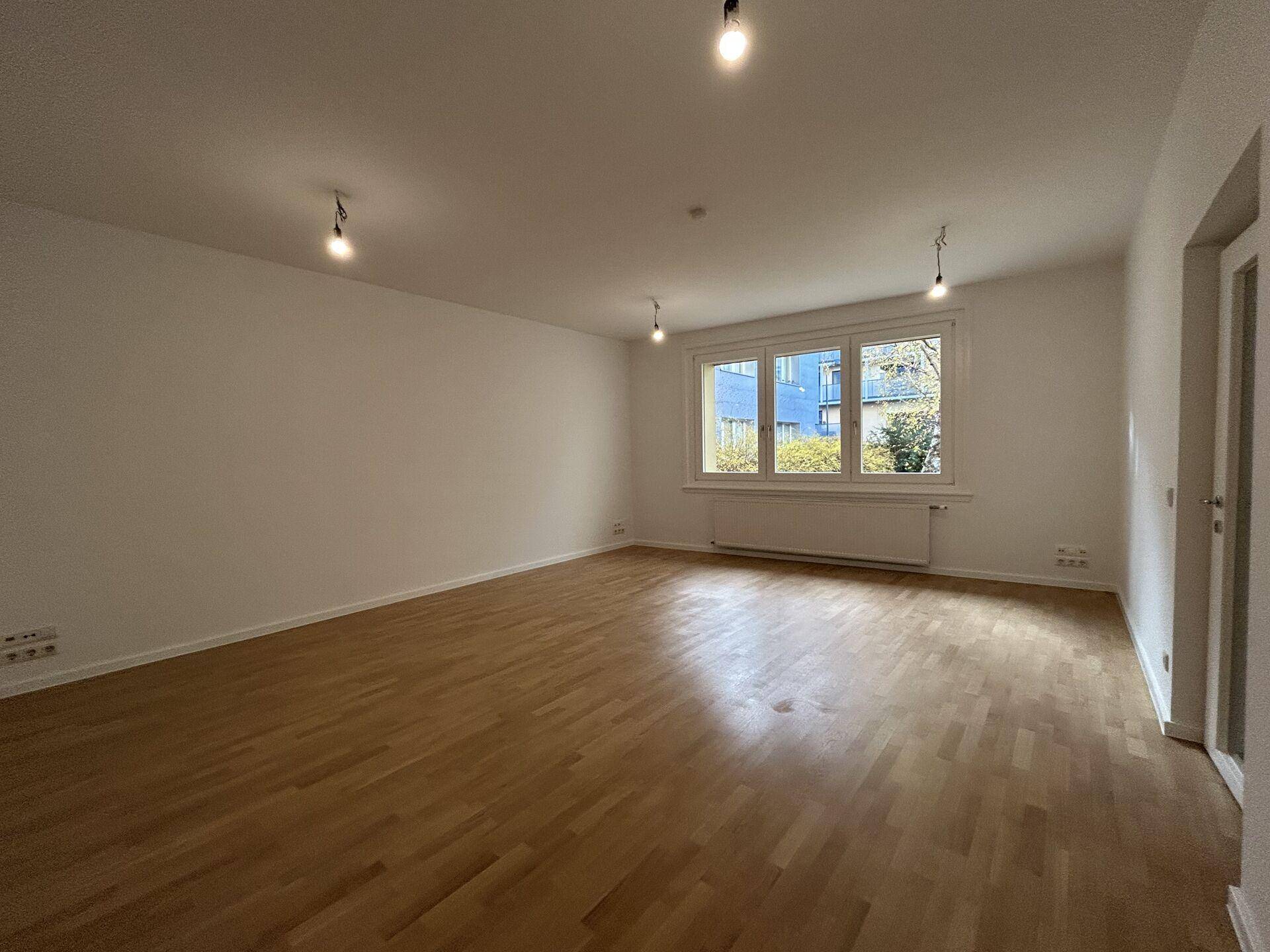 Heller, leerer Raum mit Holzboden, weißen Wänden und einem großen Fenster, das viel Tageslicht hereinlässt.