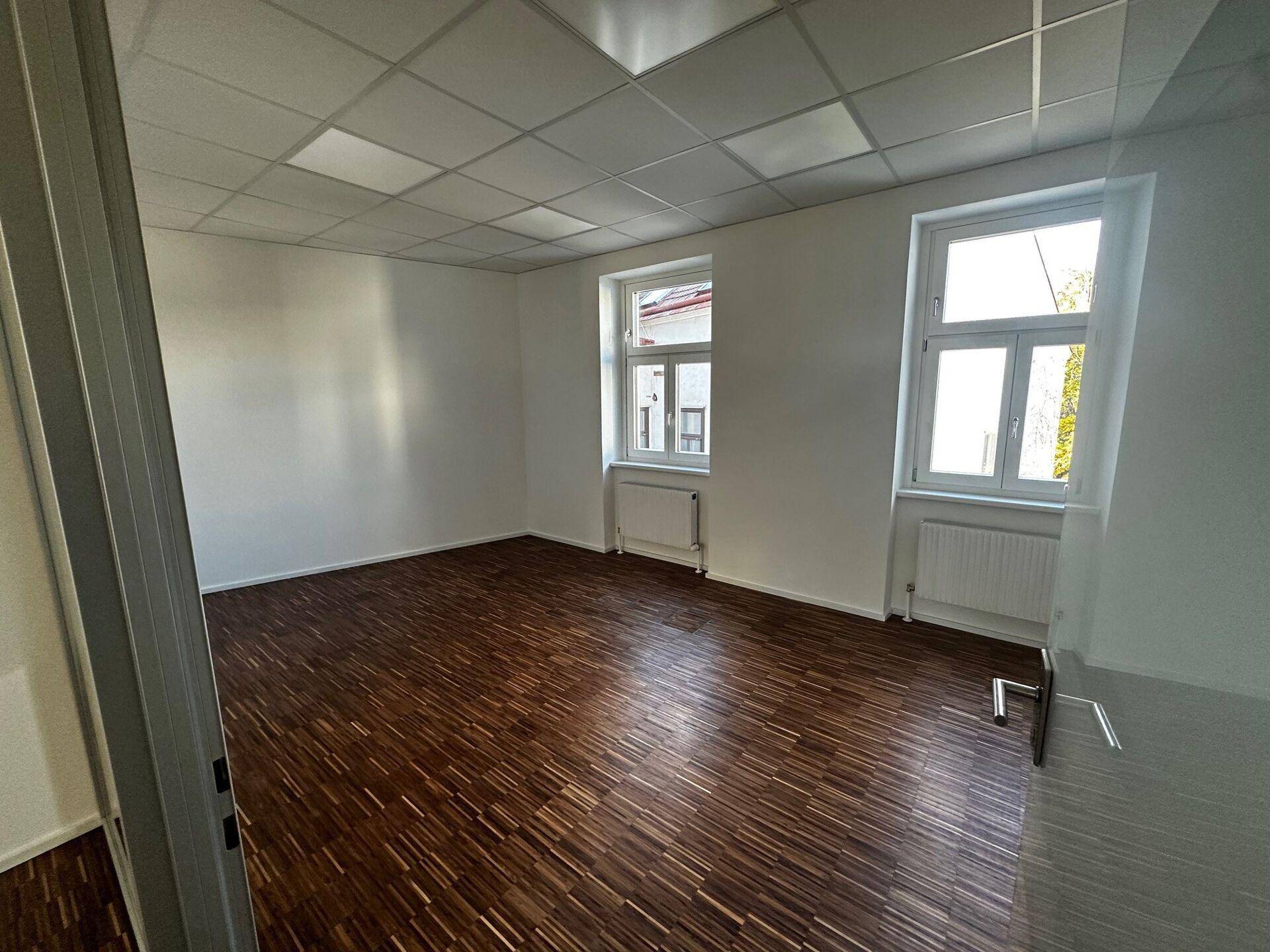 Helles, leeres Büro mit Holzboden, weißen Wänden und zwei Fenstern, die viel Tageslicht hereinlassen.