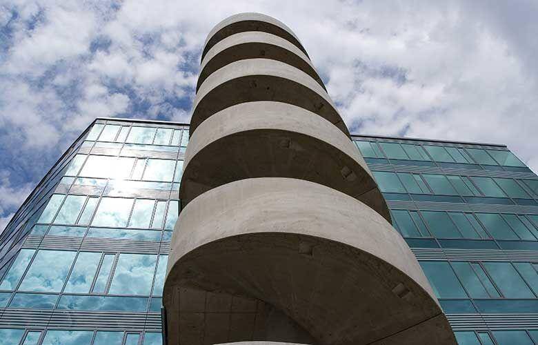 Auffällige spiralförmige Betonkonstruktion neben der modernen Glasfassade eines Bürogebäudes unter Wolken, ein architektonisches Detail.