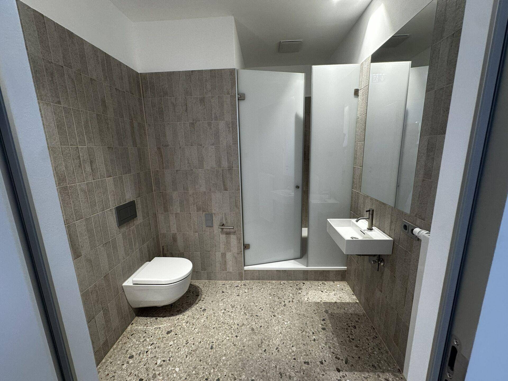 Modernes Badezimmer mit grauen Fliesen, wandhängender Toilette, begehbarer Dusche und stilvollem Waschbecken.