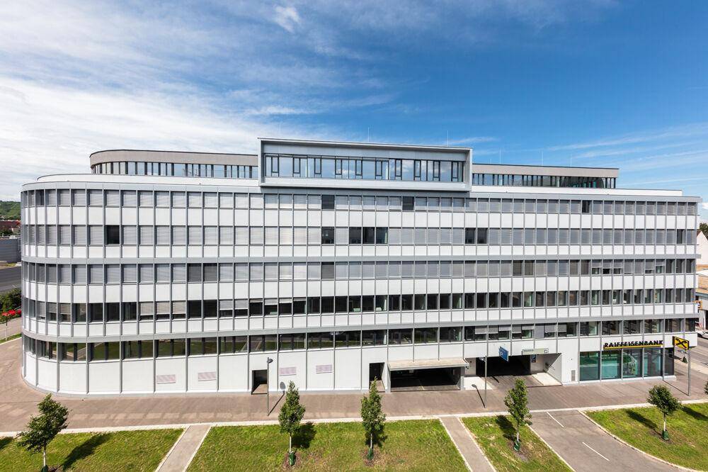 Weitläufiges modernes Bürogebäude mit vielen Fenstern und einem Parkplatzbereich im Vordergrund.