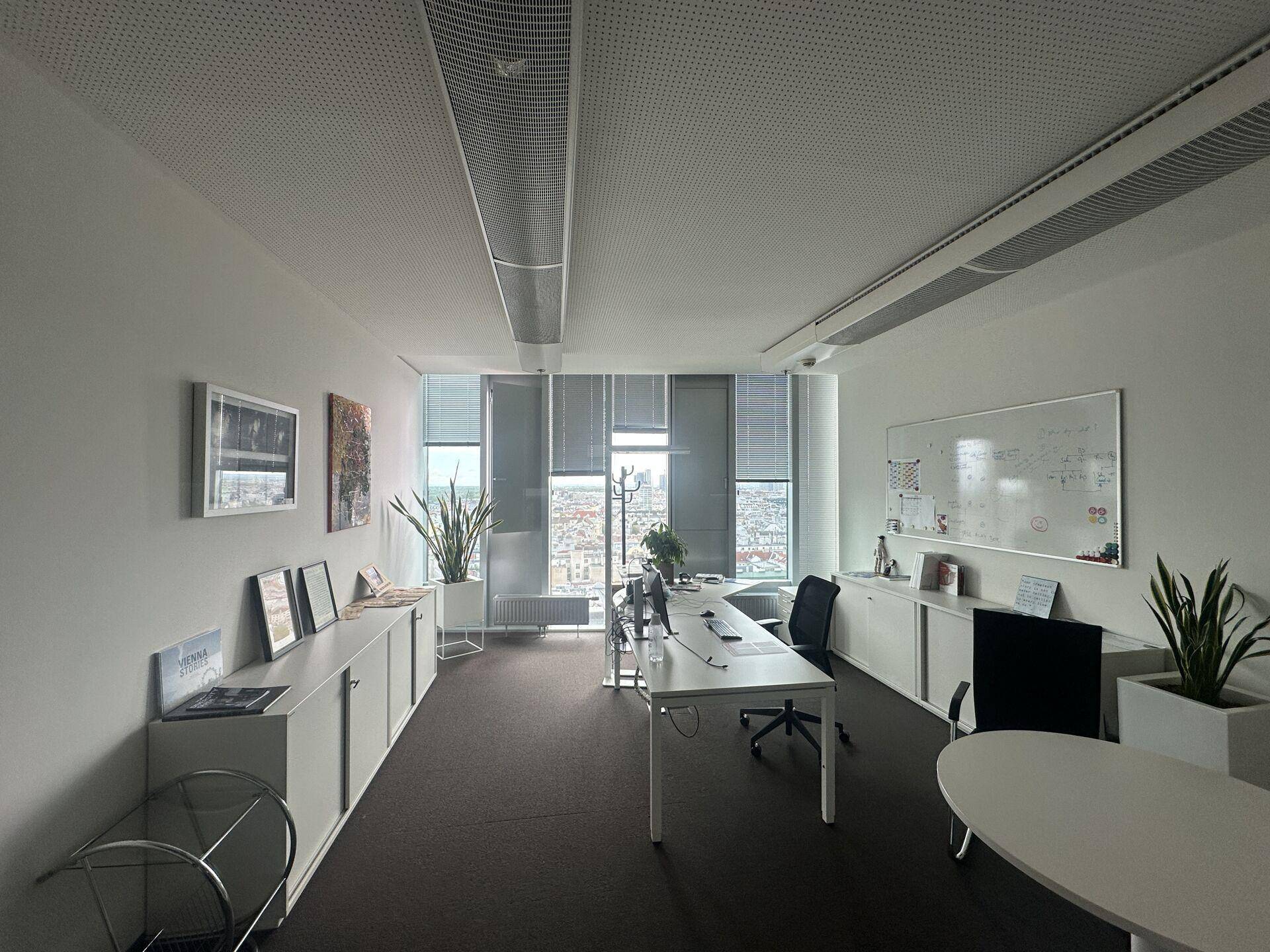 Geräumiges Büro mit langen Schreibtischen, weißen Schränken und großen Fenstern, die viel Tageslicht hereinlassen.