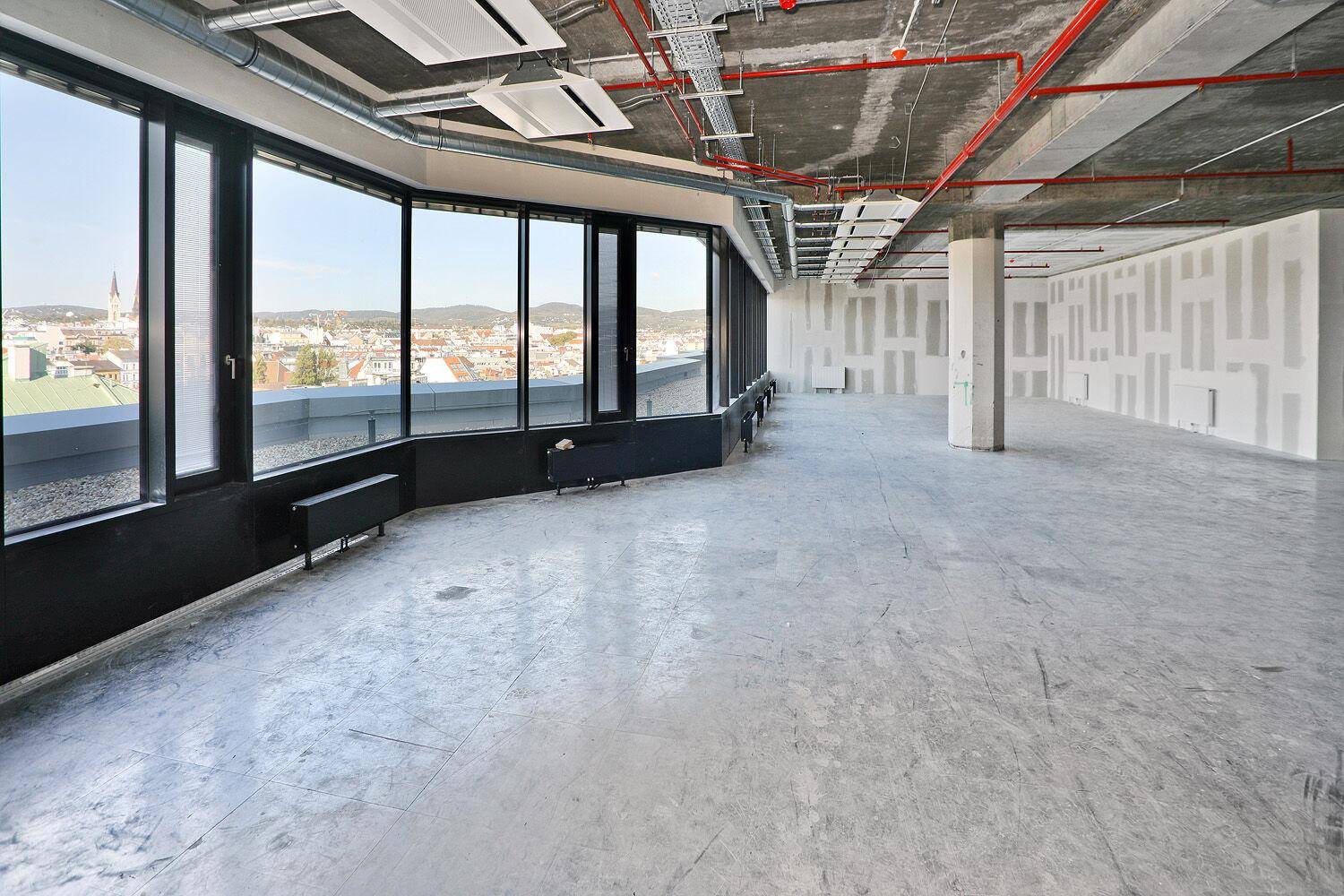 Geräumiger Büroraum im Rohbau mit unverputzten Wänden, Betonboden und großen Fenstern mit Blick auf die Stadt.
