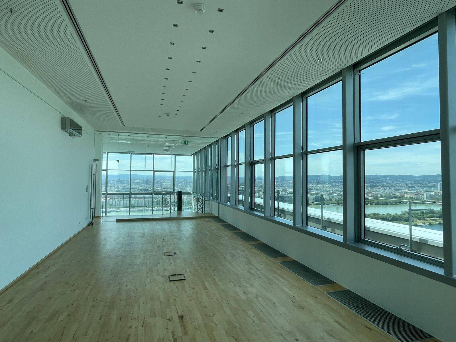 Großer, leerer Büroraum mit Holzboden und raumhohen Fenstern, die einen weiten Stadtblick bieten und viel Tageslicht hereinlassen.