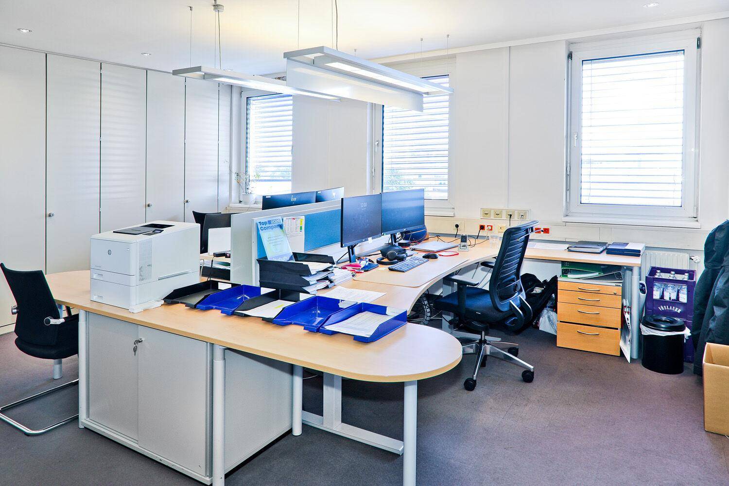 Modernes Großraumbüro mit mehreren Arbeitsplätzen, großen Fenstern und Teppichboden.